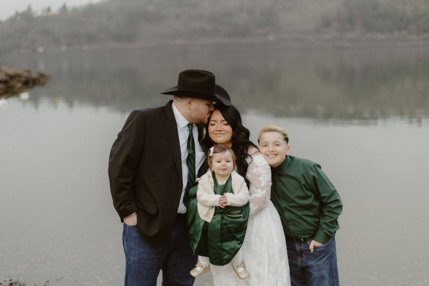 Wedding family portrait by misty lake, groom in cowboy hat, bride in lace gown with two children.