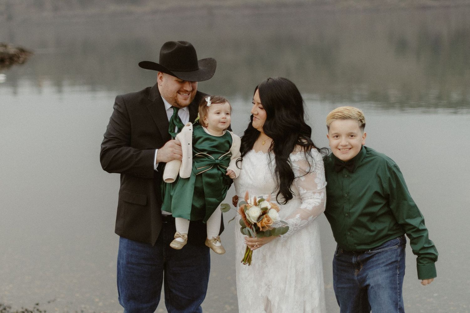 Happy wedding family portrait with groom, bride in lace dress, young daughter in green dress, and son by a lake.