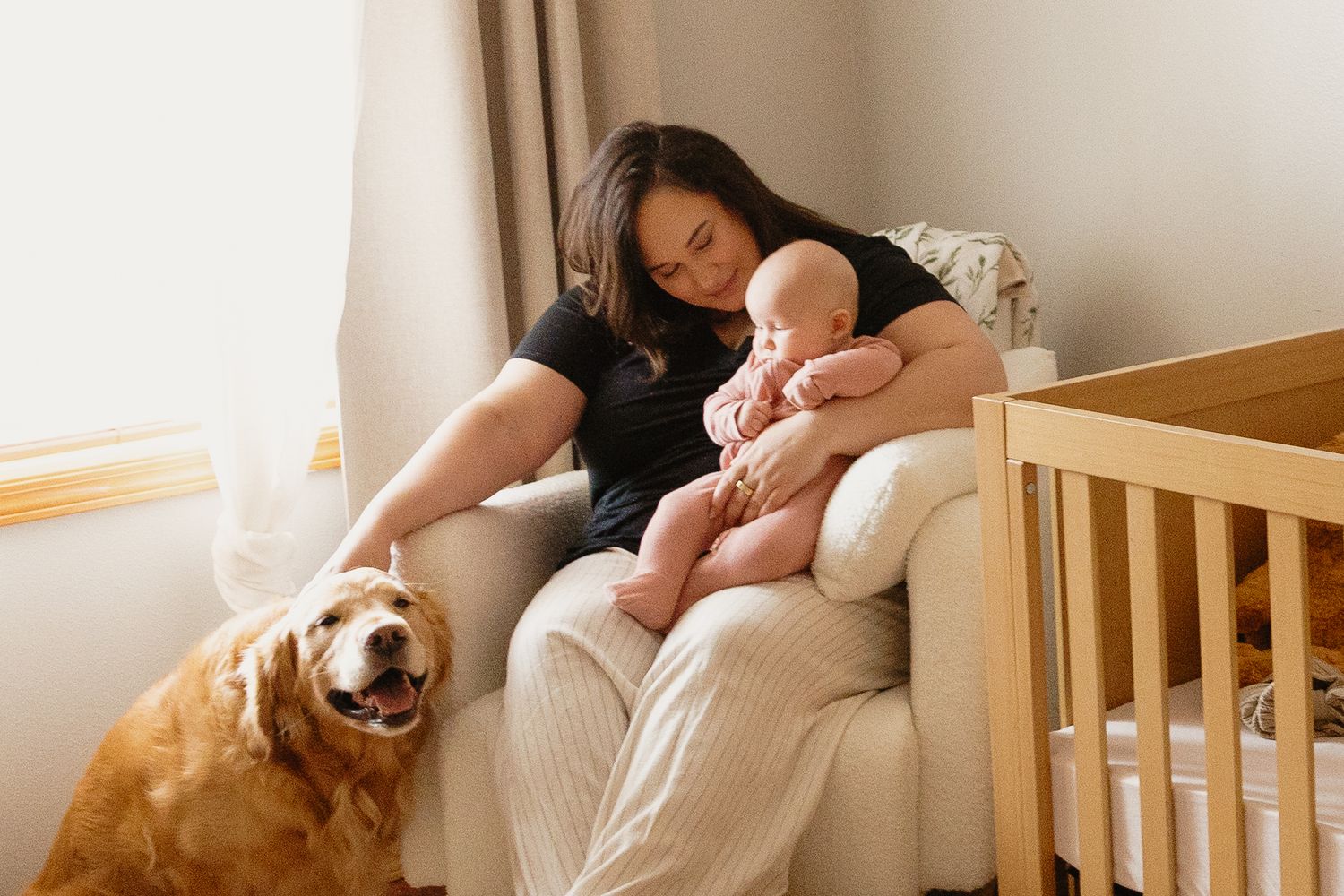 Mother sitting in nursery chair cradling newborn baby with golden retriever dog nearby.