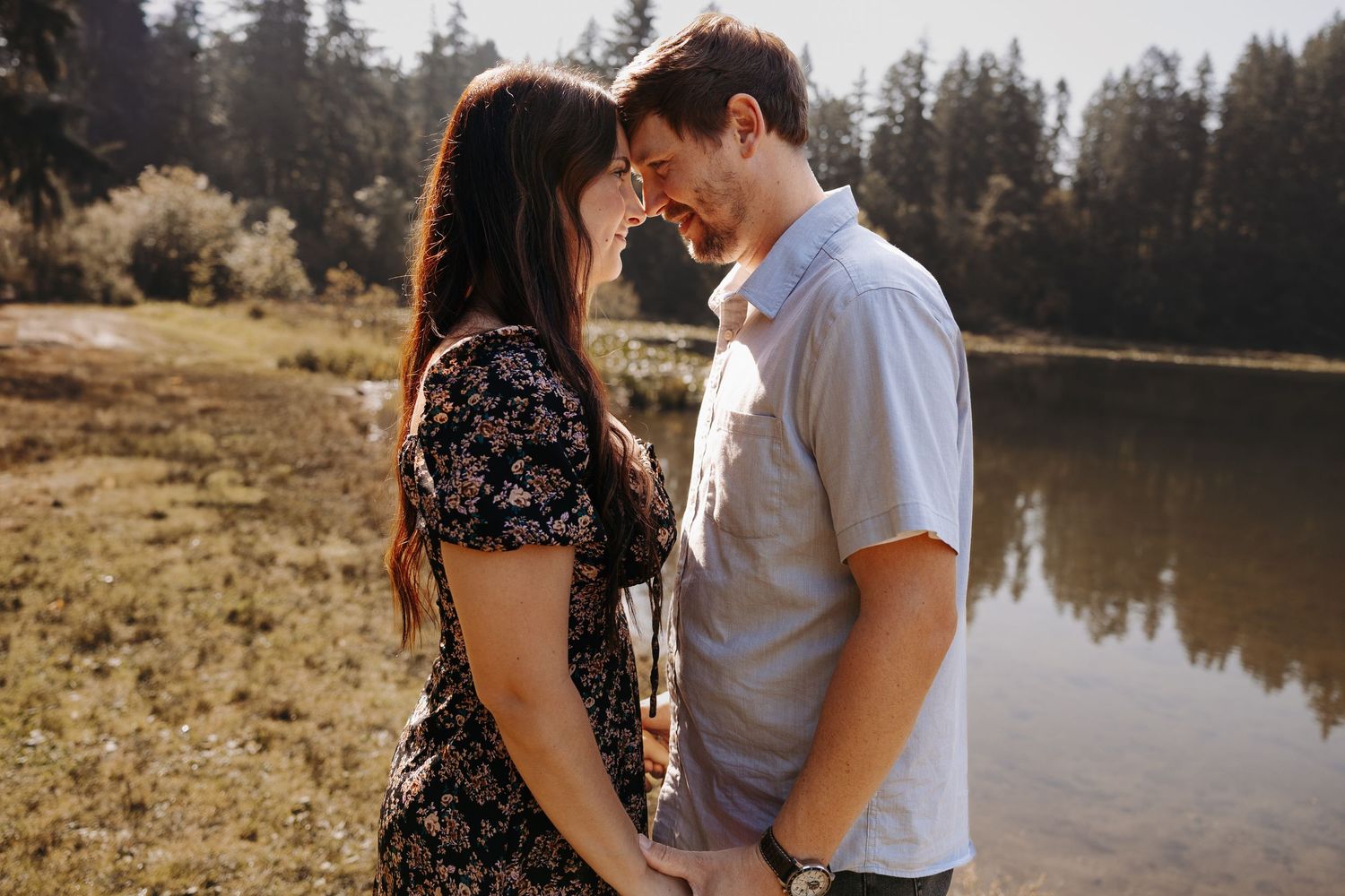A romantic couple holds hands and touches foreheads tenderly beside a sunlit lakeside setting.