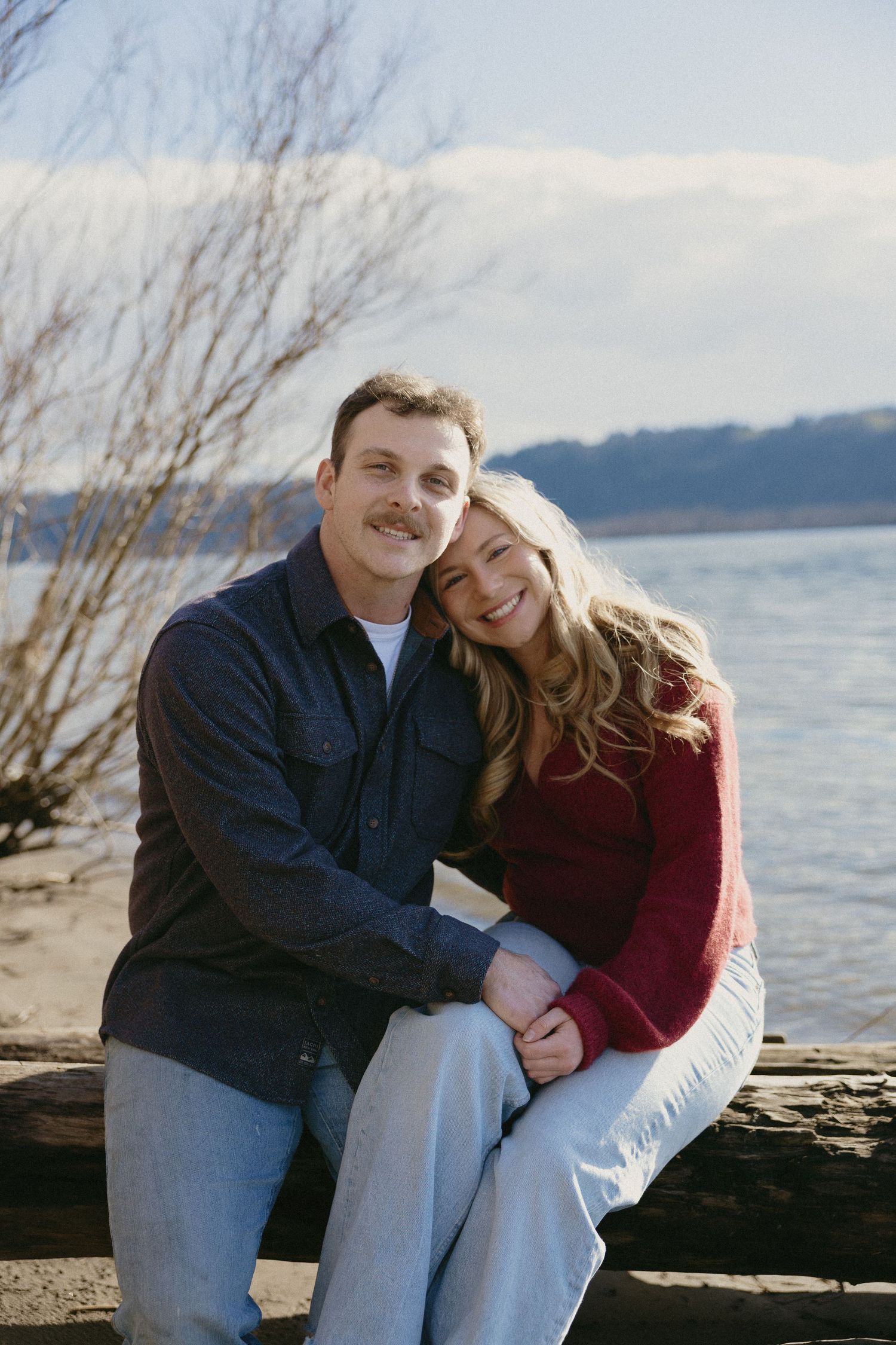 Smiling couple sitting on driftwood by a lake shore, woman leaning on mans shoulder affectionately.