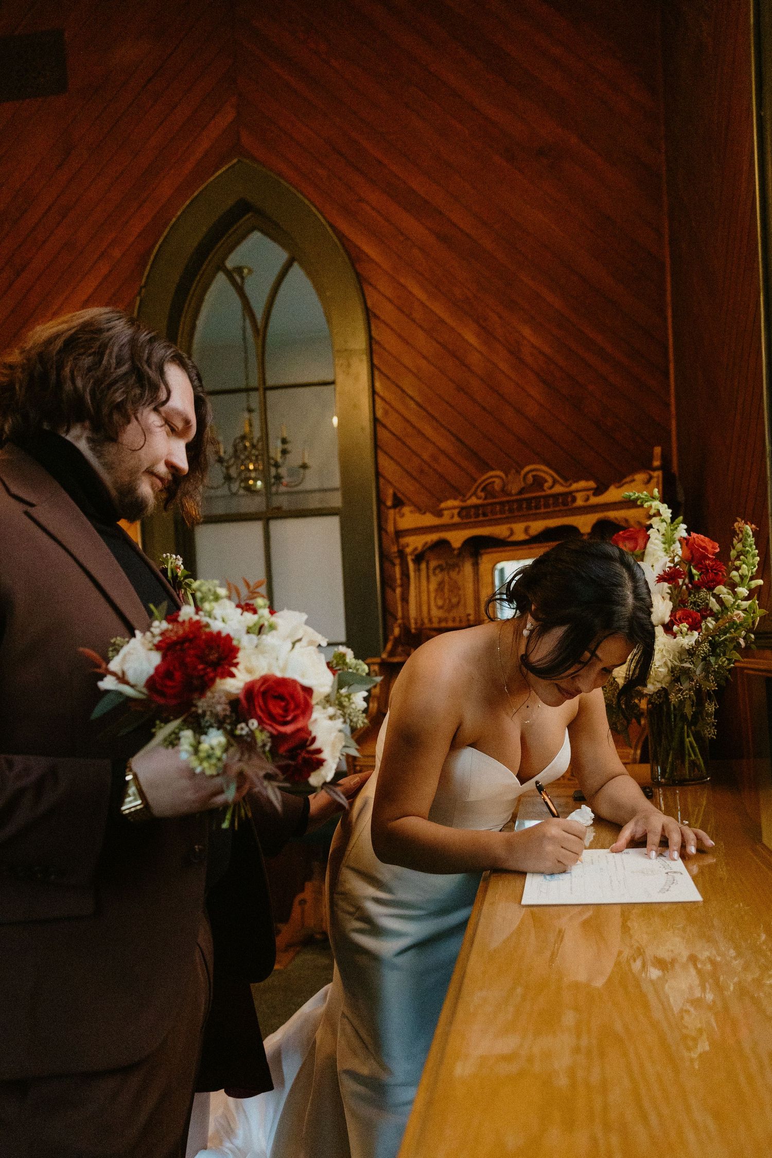 Bride signing marriage certificate while groom holds red and white bouquet in a wooden chapel.