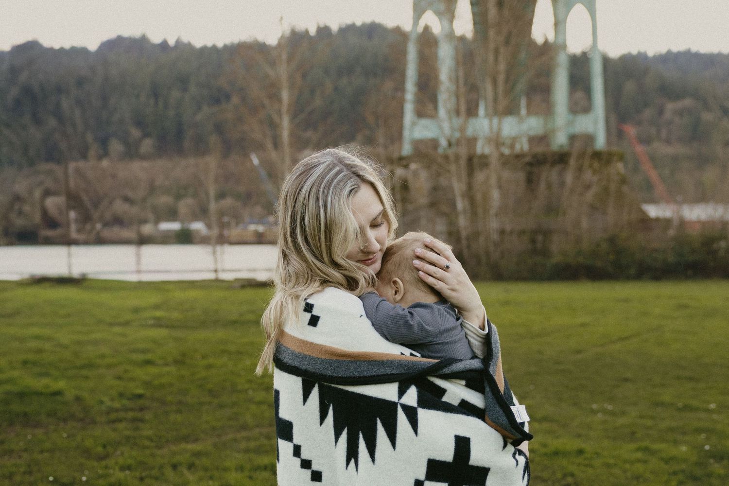 Blonde mother wrapped in Pendleton blanket cradling infant outdoors near St. Johns Bridge in Portland Oregon.