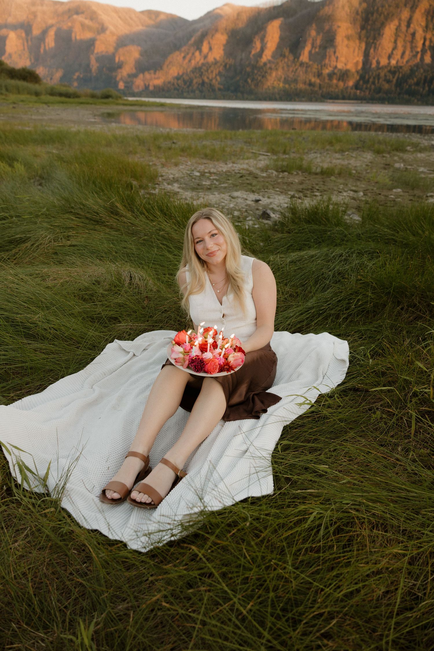 Young blonde woman sitting on blanket in grass holding floral birthday cake at sunset.