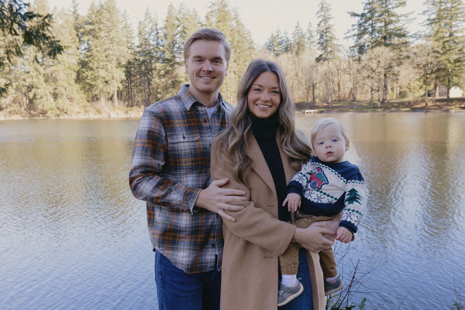 Young family of three smiling together by a calm lake with evergreen trees in autumn sunlight.