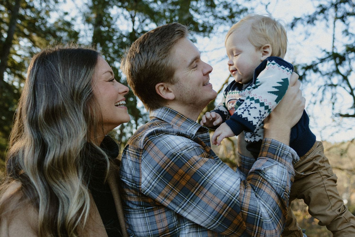 Smiling young parents in woodland setting, father lifting laughing toddler in holiday sweater.