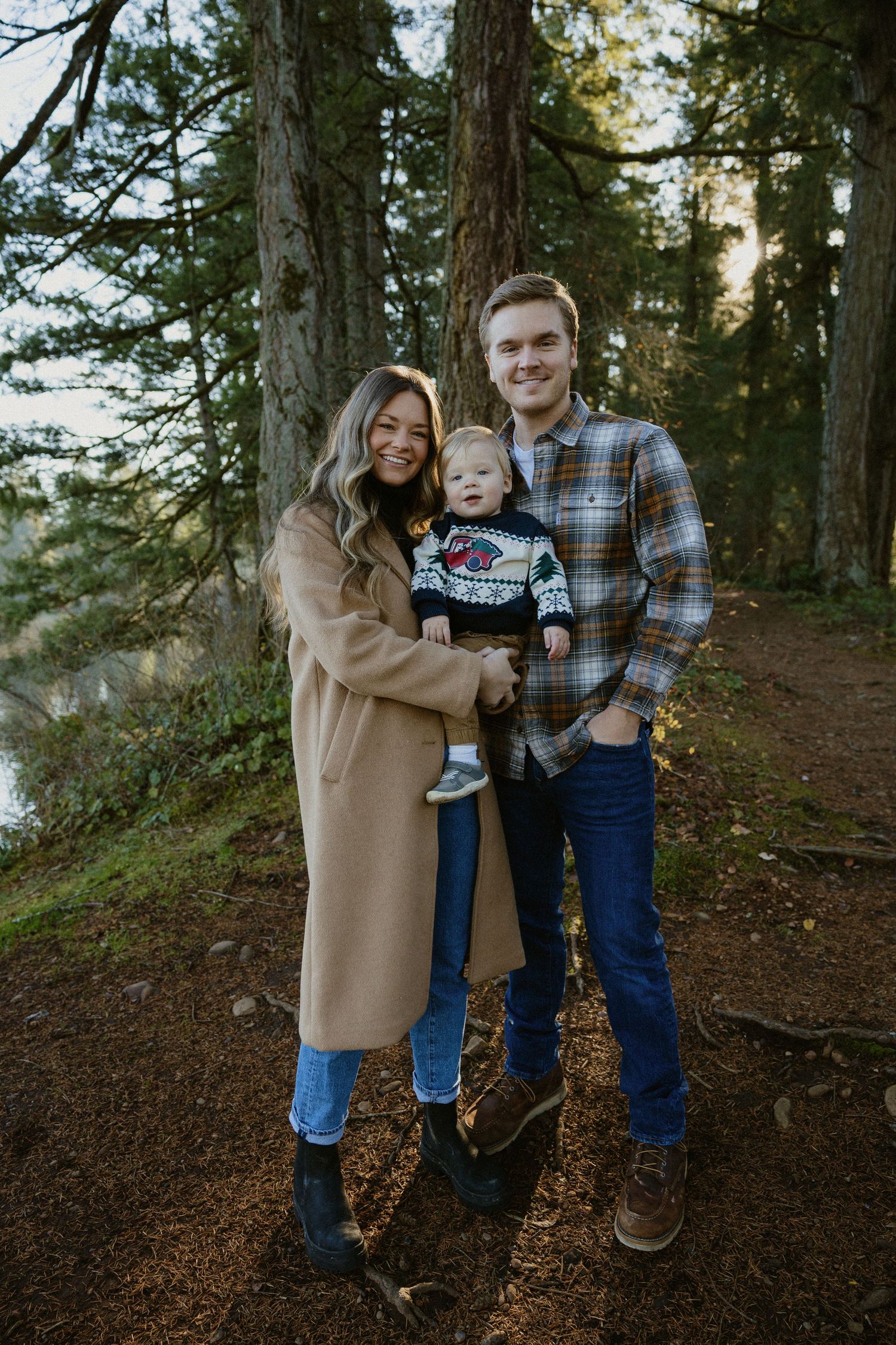 Young family with baby boy posing in forest wearing flannel and camel coat for fall photos.