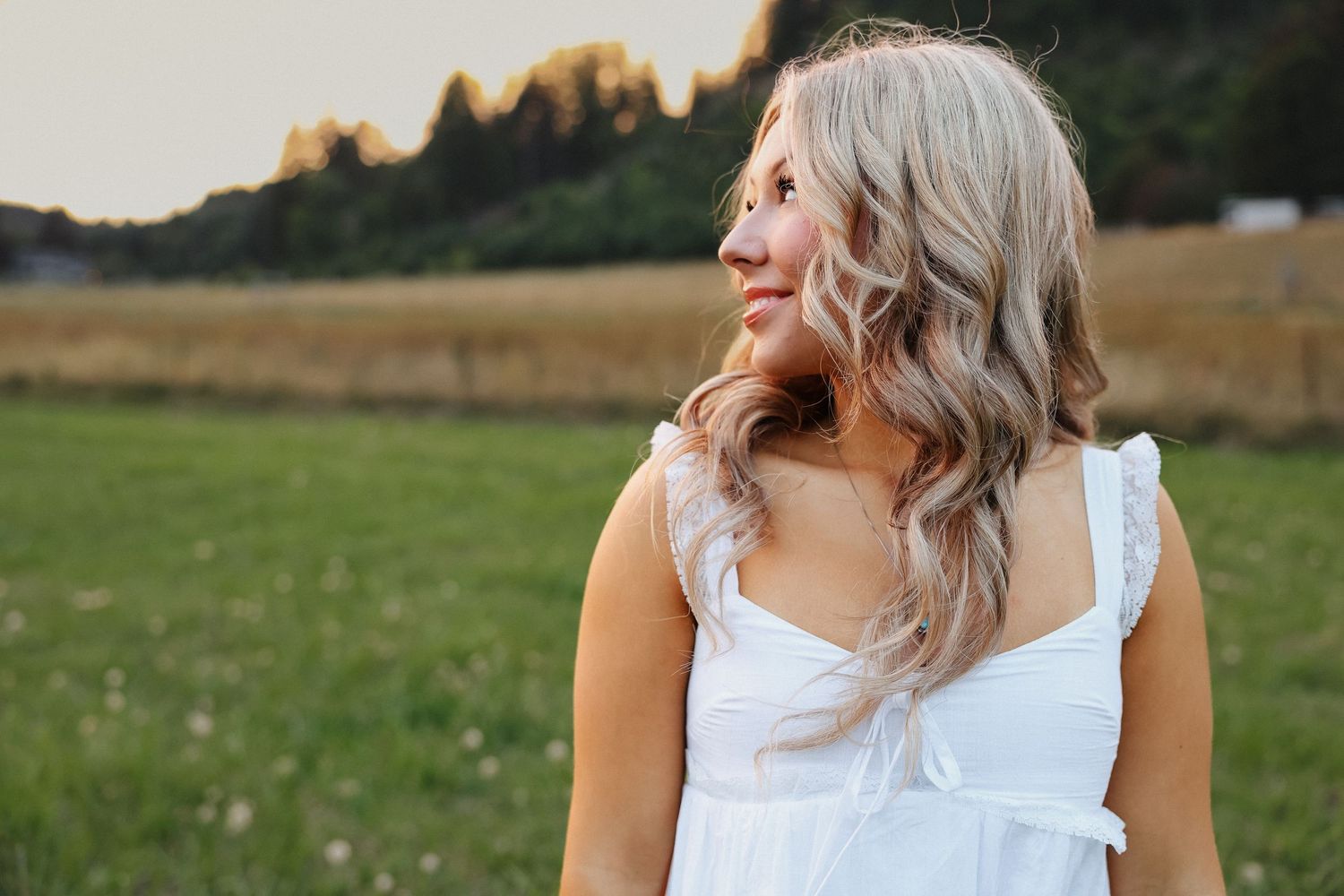 Blonde woman in white sundress looking away in green field at golden hour sunset.
