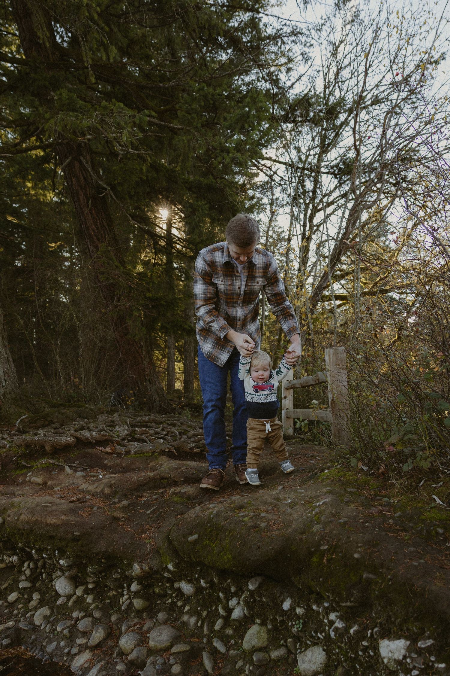 A father helps his toddler walk on rocky terrain in a forest setting during autumn.