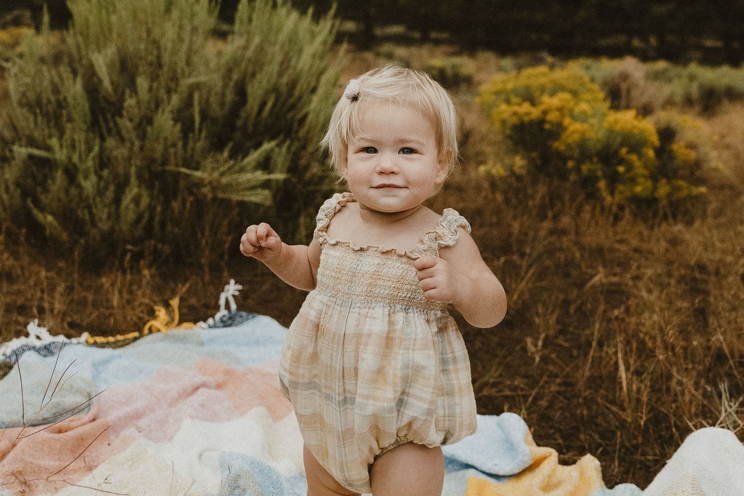 Smiling baby girl in plaid romper sitting on colorful blanket in golden field.