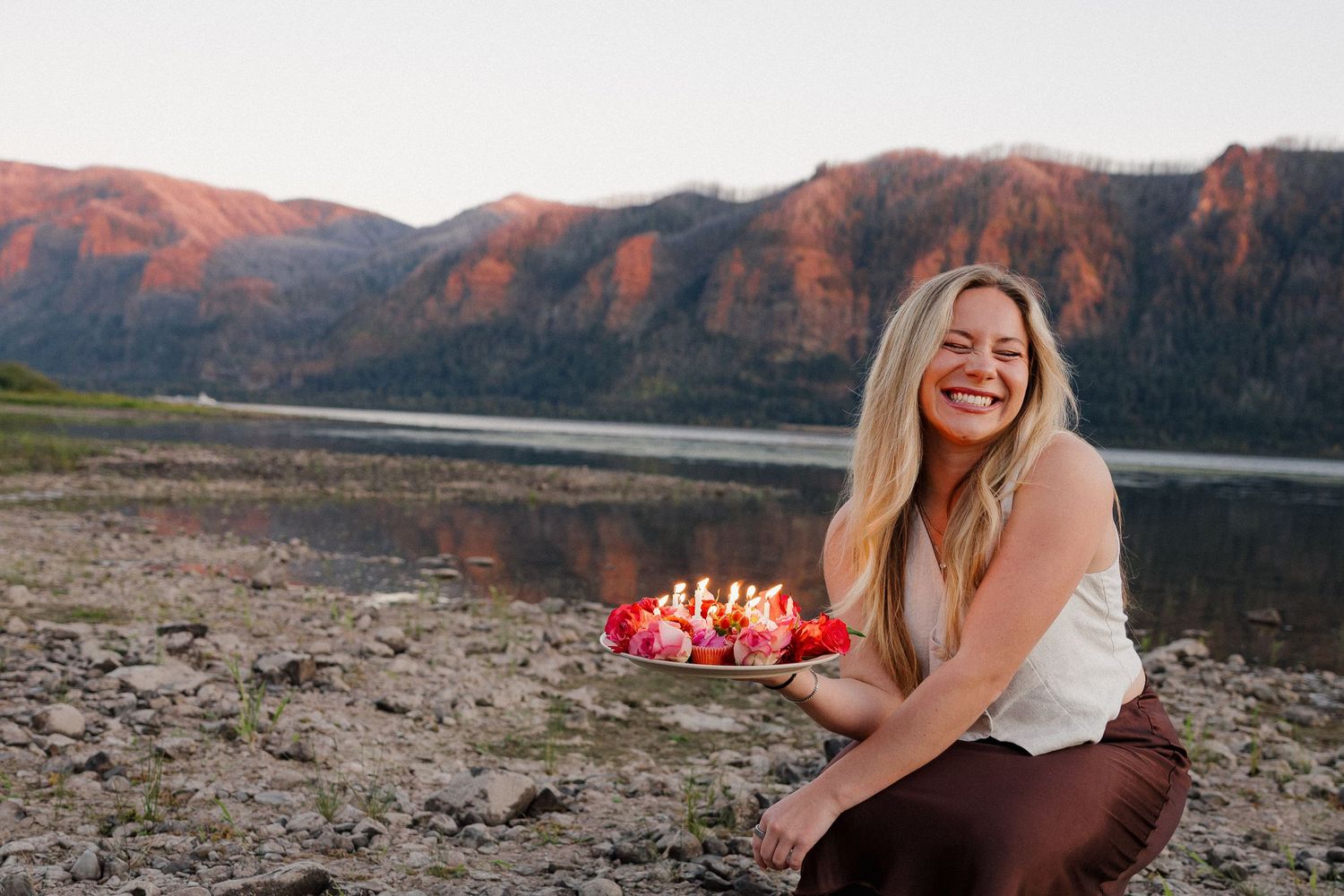 Laughing blonde woman crouching by mountain lake holding a floral birthday cake at dusk.