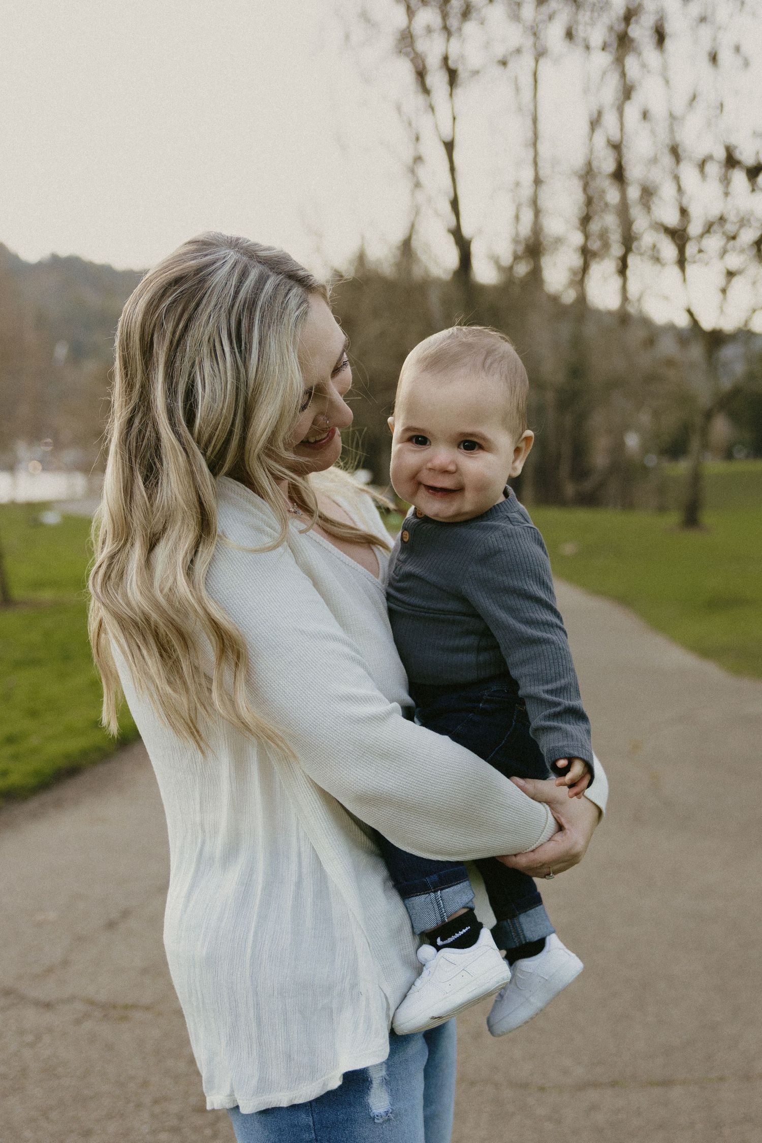 Smiling blonde mother in white sweater holding laughing baby boy on park pathway in Portland Oregon.