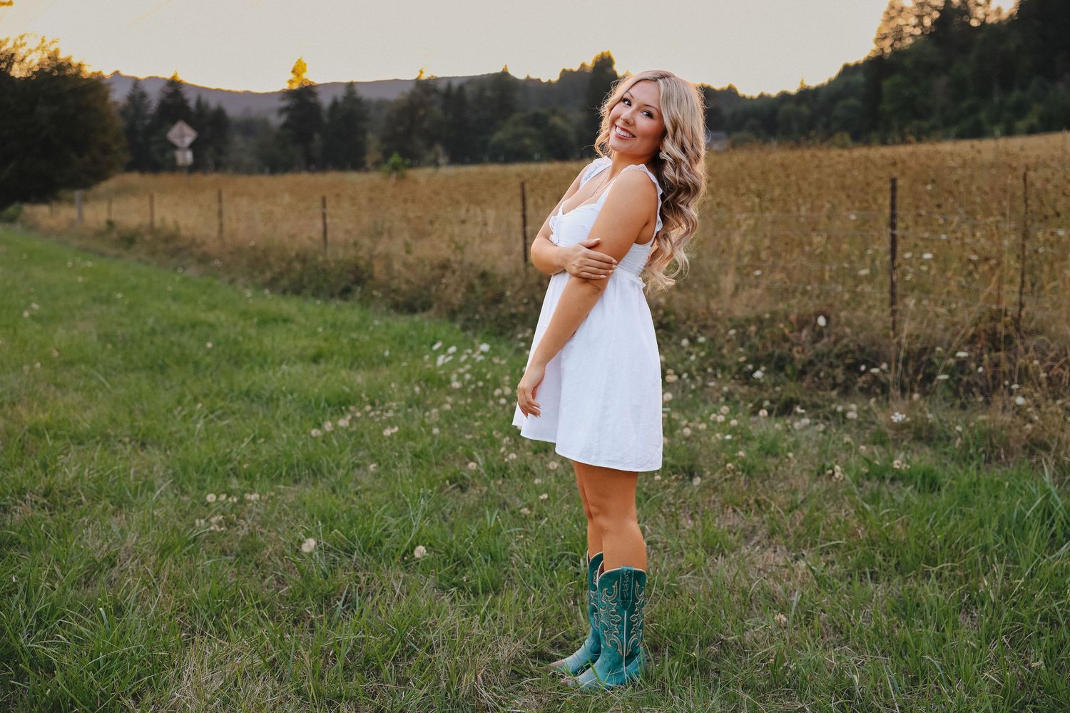 Blonde woman in white dress and turquoise cowboy boots posing in green field at sunset.