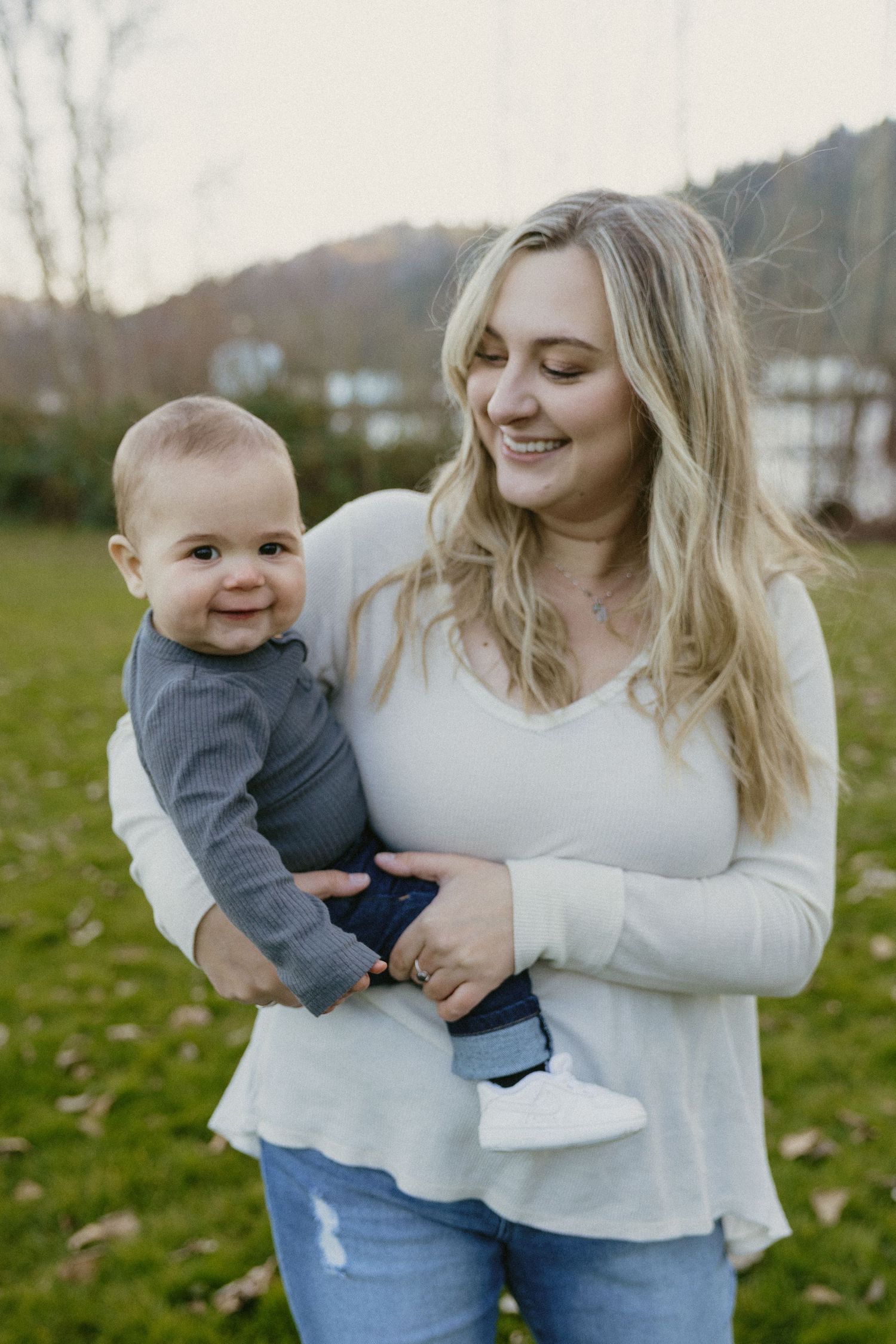 Smiling blonde mother in white top holding happy baby boy in gray outfit outdoors.