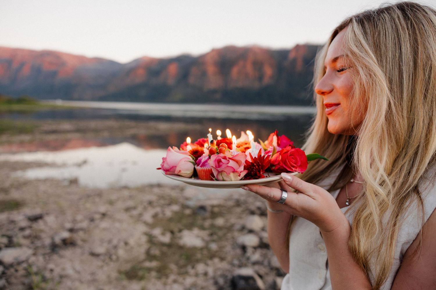 Blonde woman smelling a floral birthday cake with lit candles at golden hour by a lake.