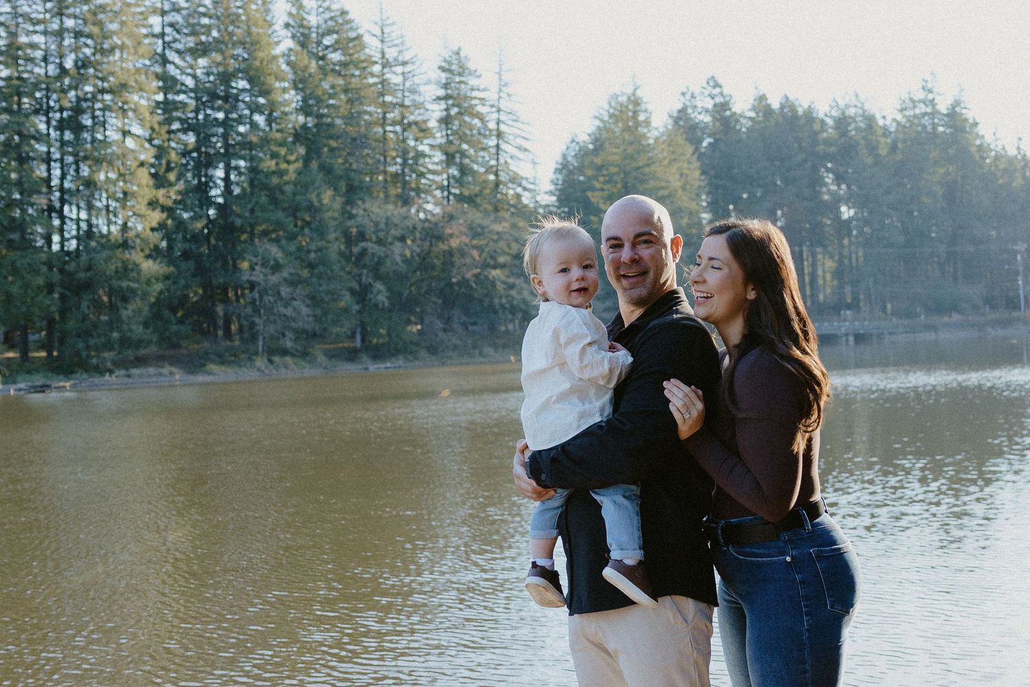 Joyful family of three laughing together outdoors by a sunlit lake surrounded by evergreen trees.