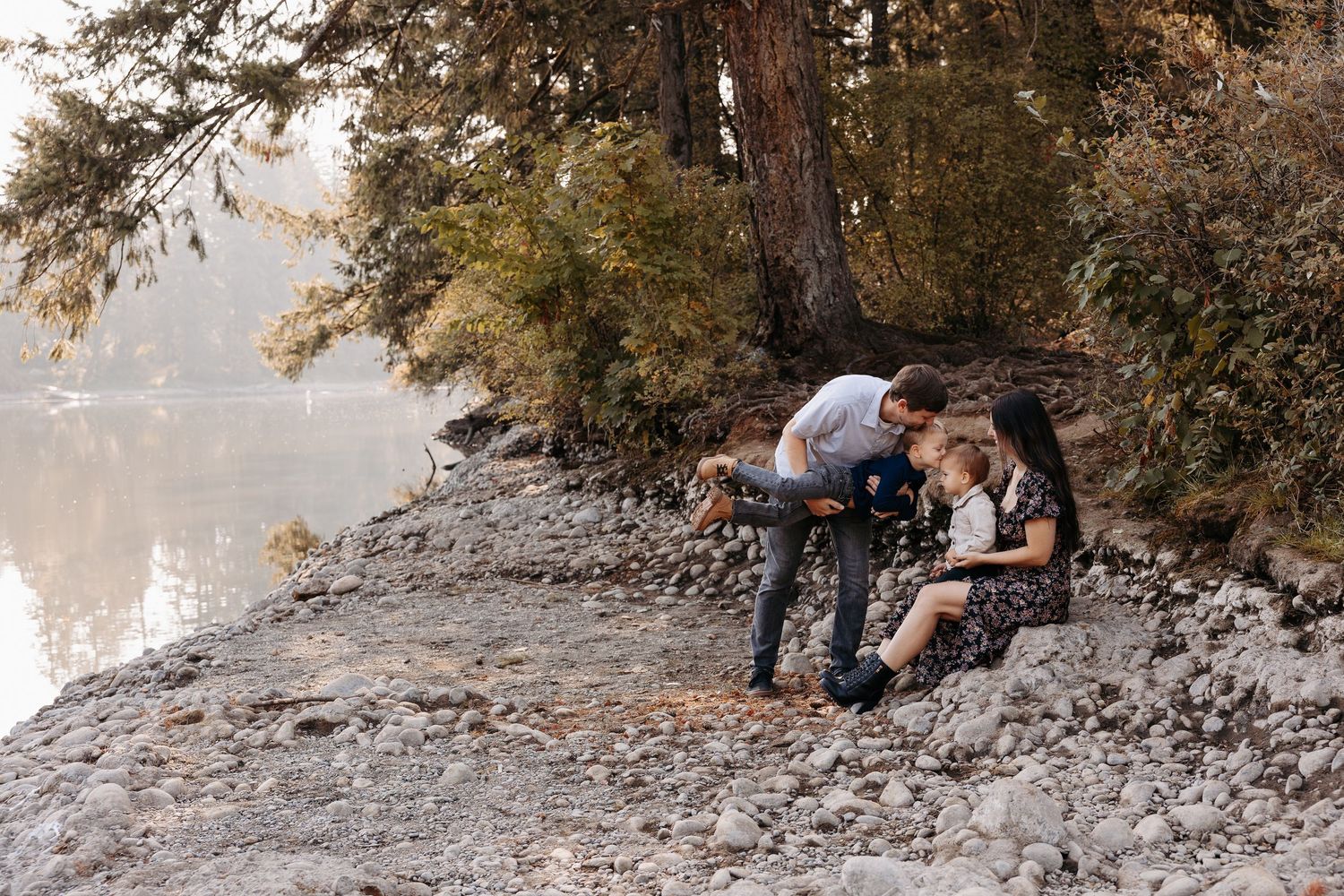 Family exploring rocky lakeside shore together, parents and two young children near calm water.