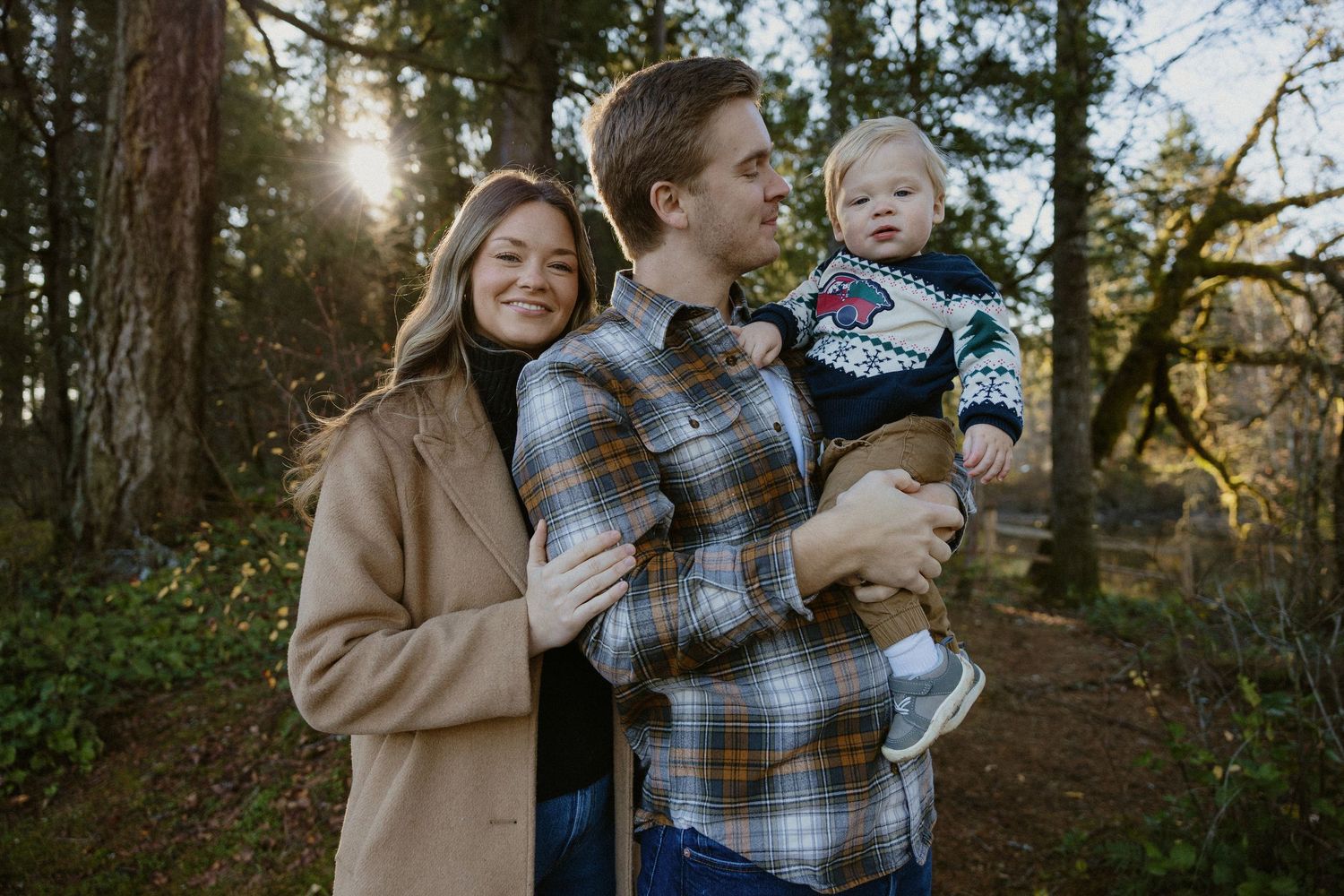 Young couple smiling outdoors in forest, man holding toddler in festive sweater at sunset.