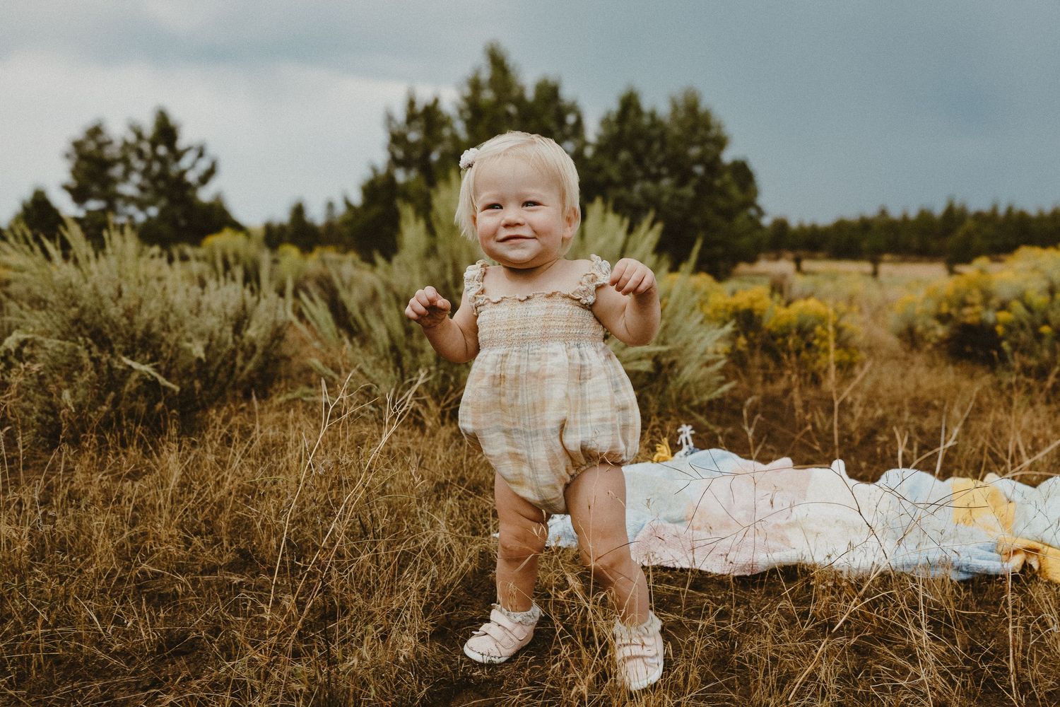 Happy baby girl in plaid romper stands smiling in golden meadow with colorful blanket nearby.