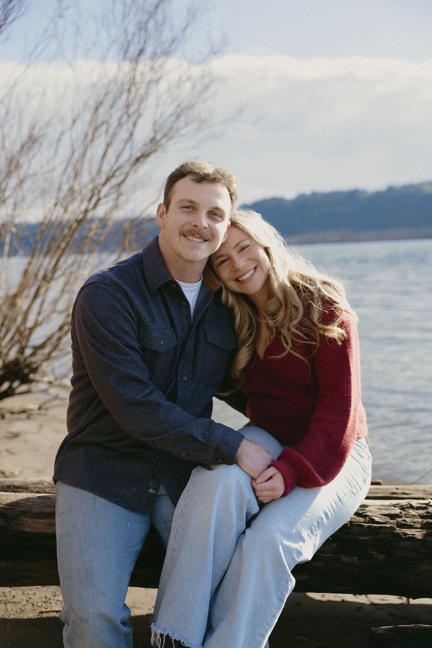 Happy couple sitting together on driftwood log with lake and winter trees in background.