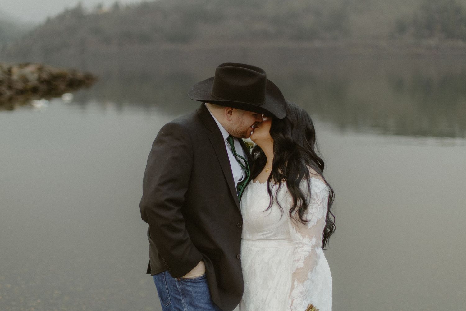 Bride and groom lean in for a kiss outdoors near a serene misty lake, groom wearing a black cowboy hat.