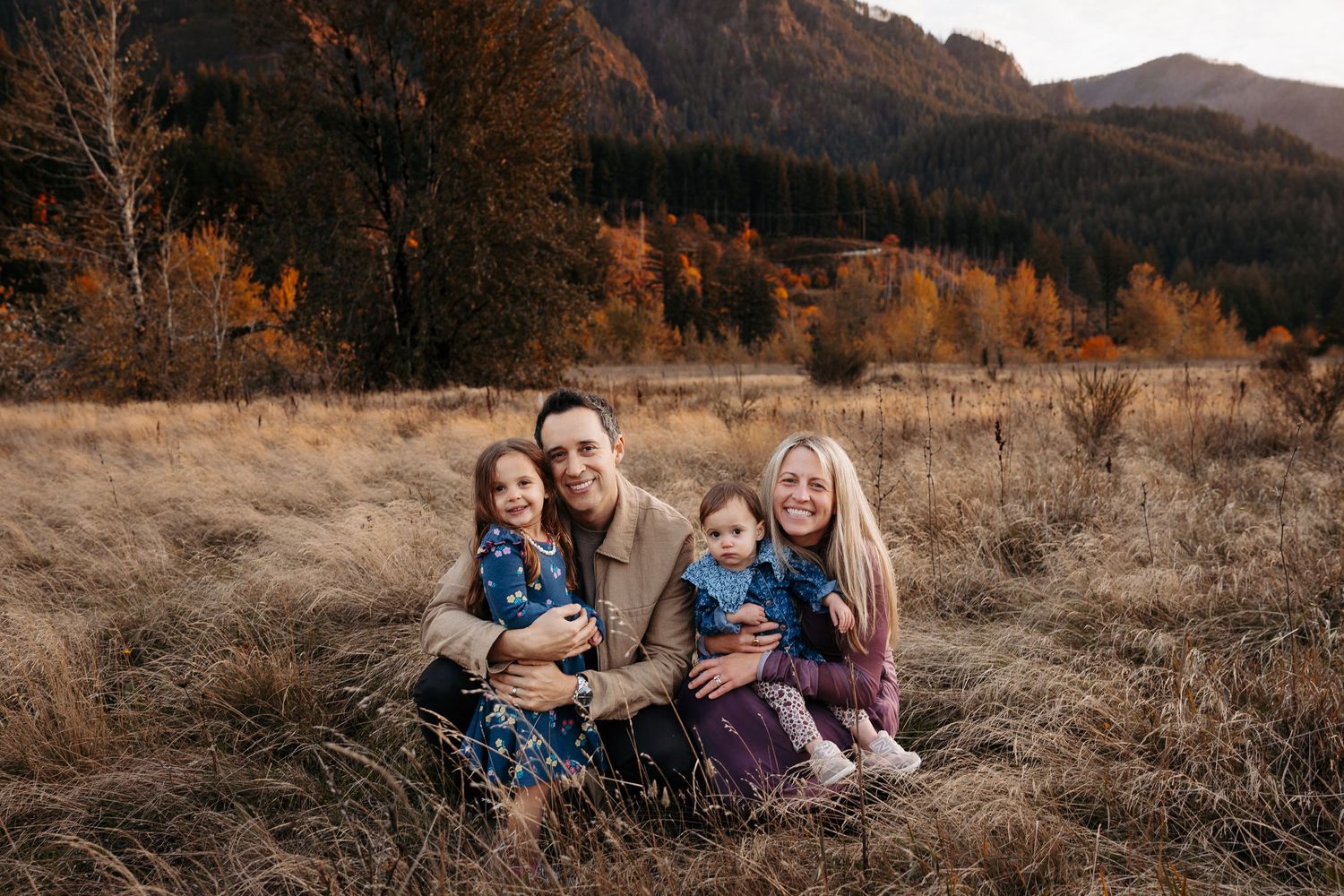 Smiling family of four sitting together in golden meadow with autumn foliage and mountains behind.