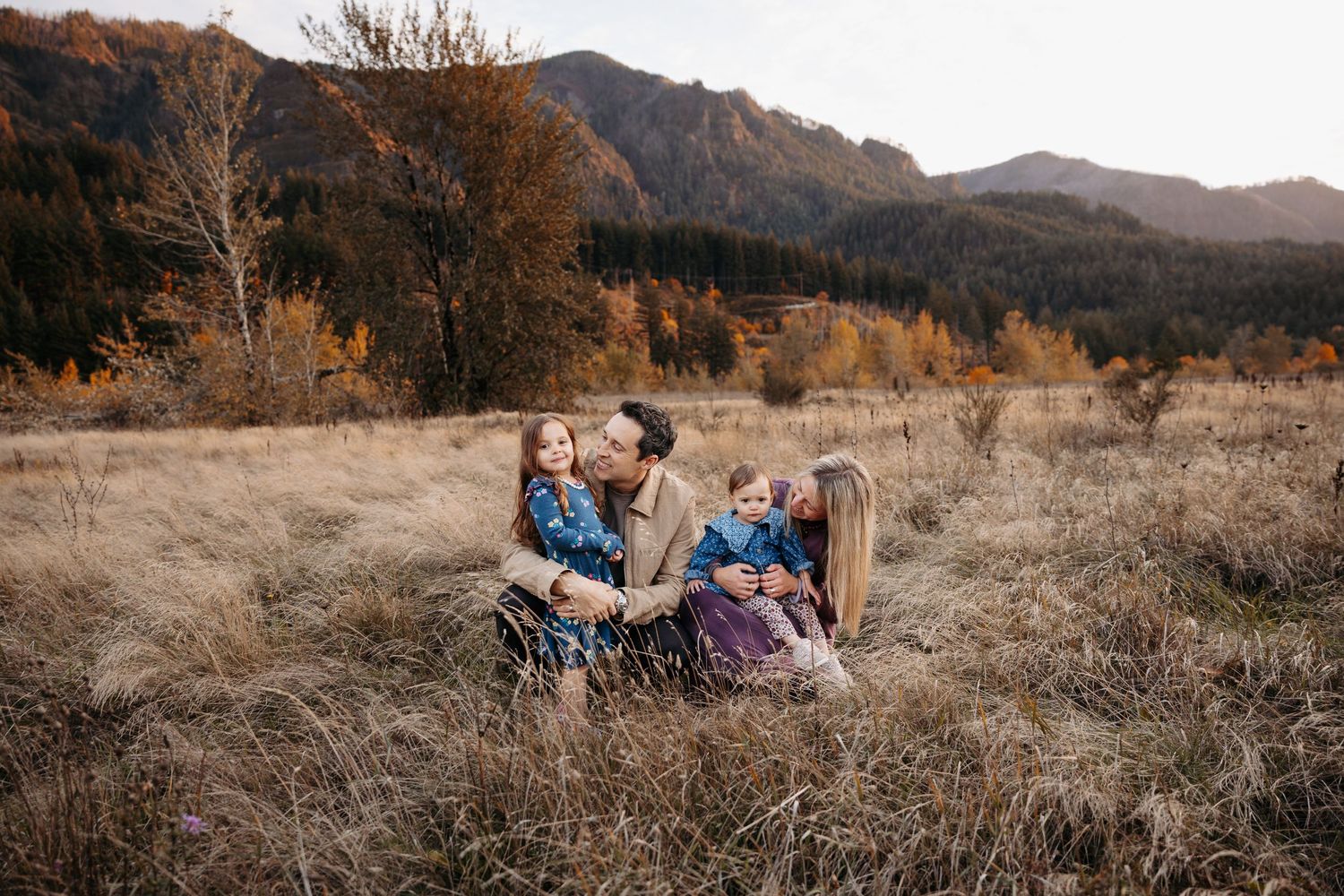 Family of four sitting in golden autumn meadow with mountain backdrop during fall season.