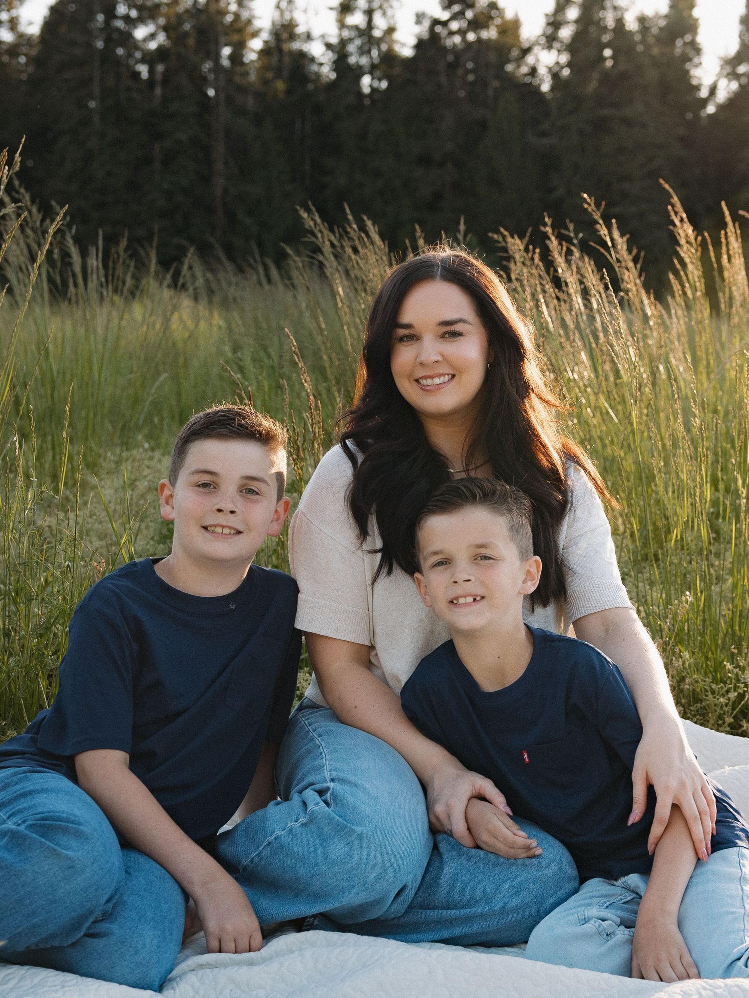 A smiling woman poses with two young boys on a blanket in a sunlit field with tall grass behind them.