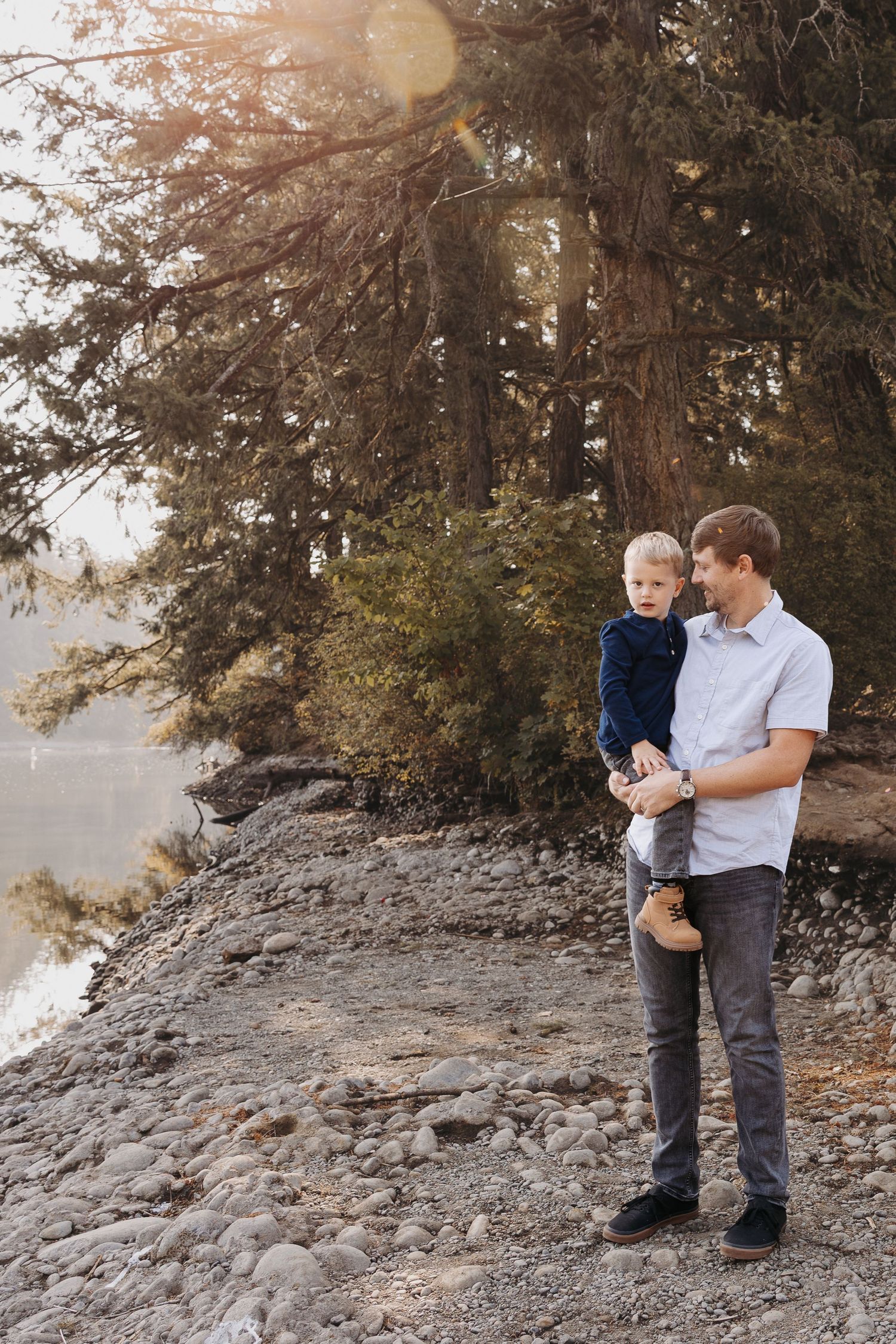 Father holding young toddler son near rocky lakeside surrounded by tall evergreen trees.
