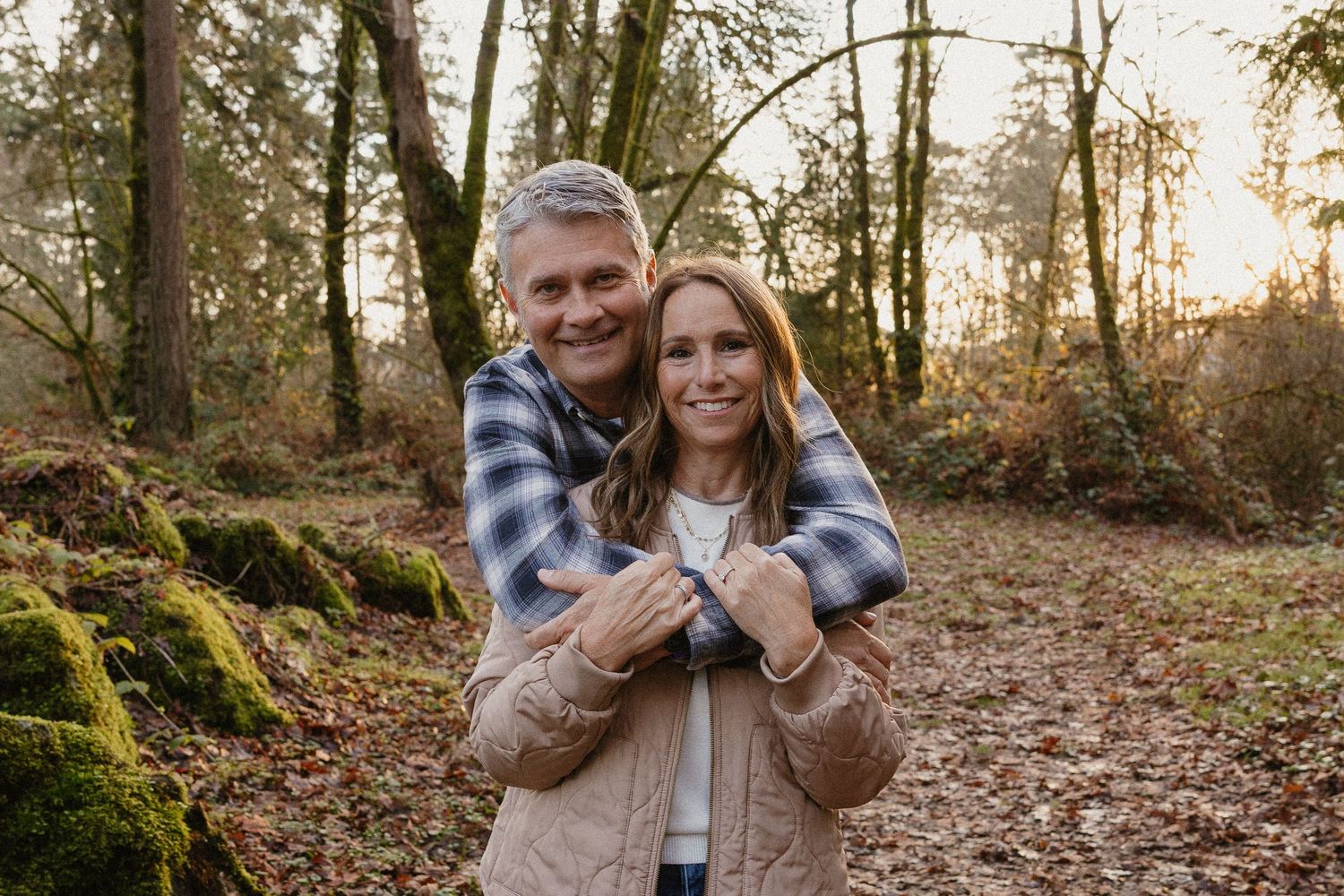 A smiling couple embraces on a leaf-covered forest path during golden hour.