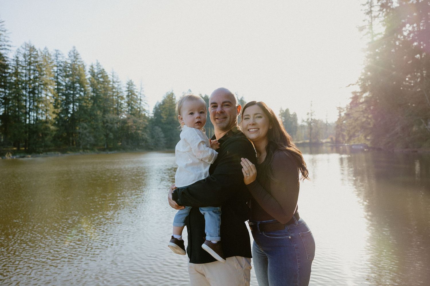 Happy family of three posing with a baby by a glowing sunlit lake and forest backdrop.