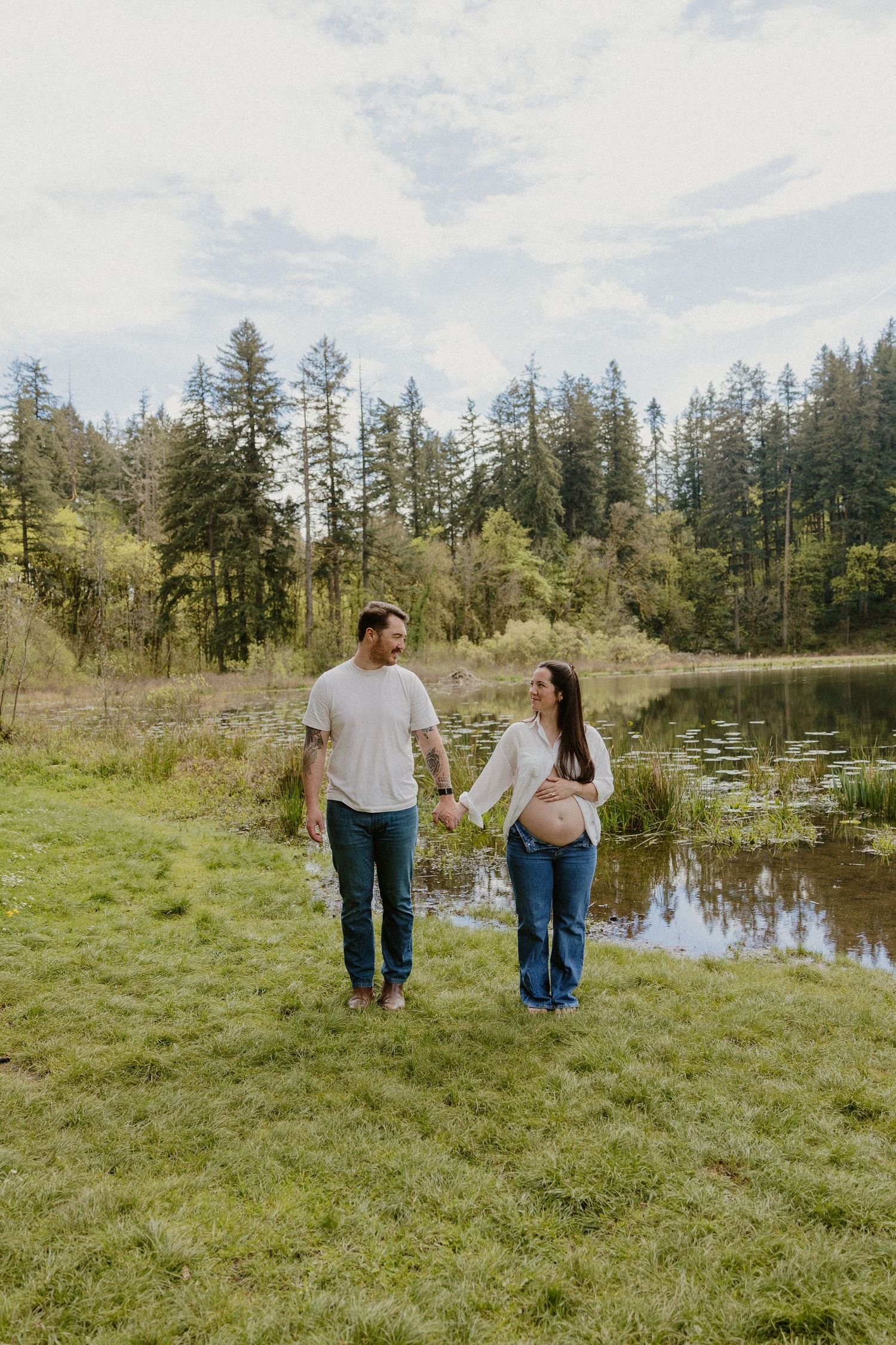 Happy expecting couple holding hands and walking along a scenic pond with tall evergreen trees.