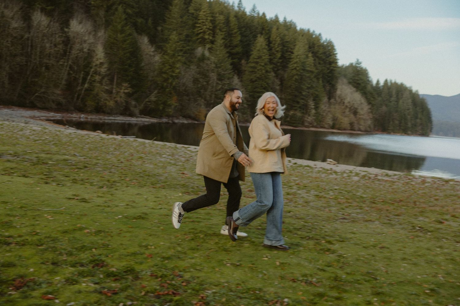 Joyful couple running hand in hand along a lakeside with forest backdrop.