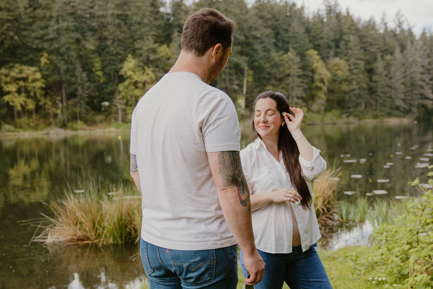 Couple smiling and holding hands near a peaceful forest pond during a maternity photo session.