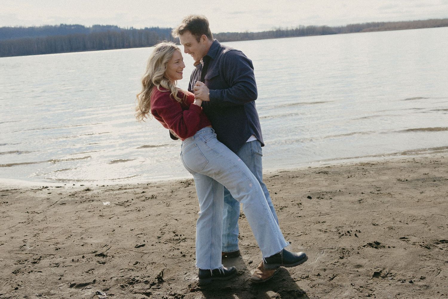 Couple embracing and smiling at each other on a sandy beach by a calm lake in fall.