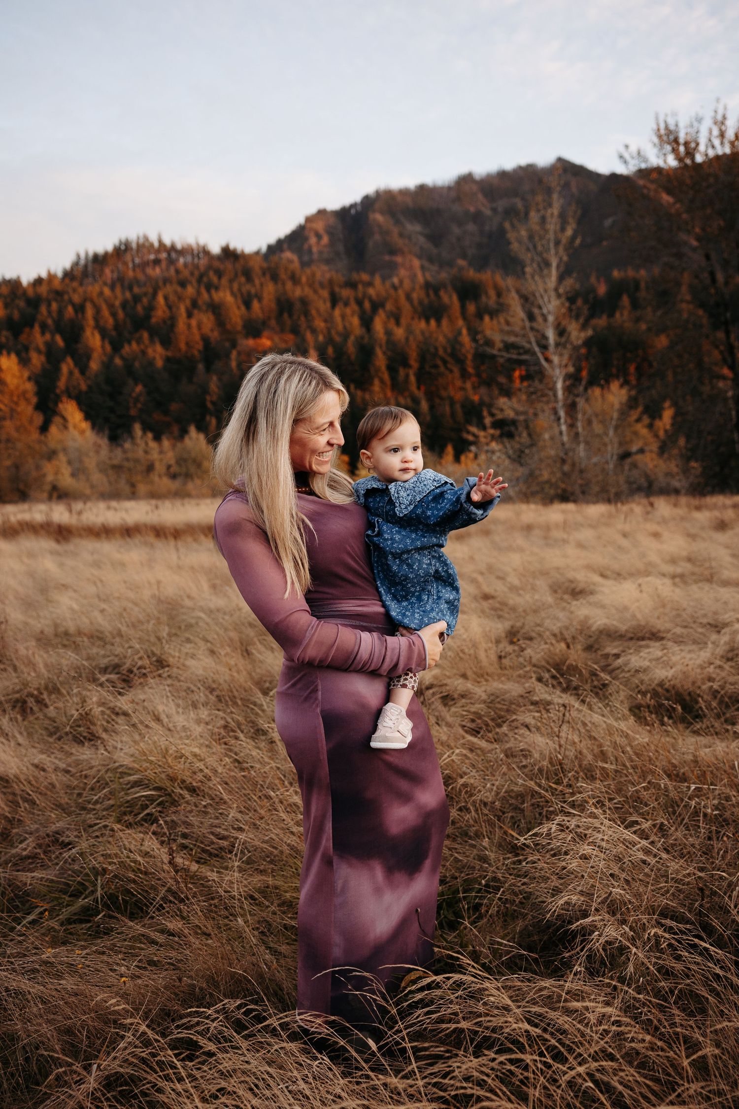 A blonde mother in a purple dress holds her curious baby outdoors in a fall mountain setting.