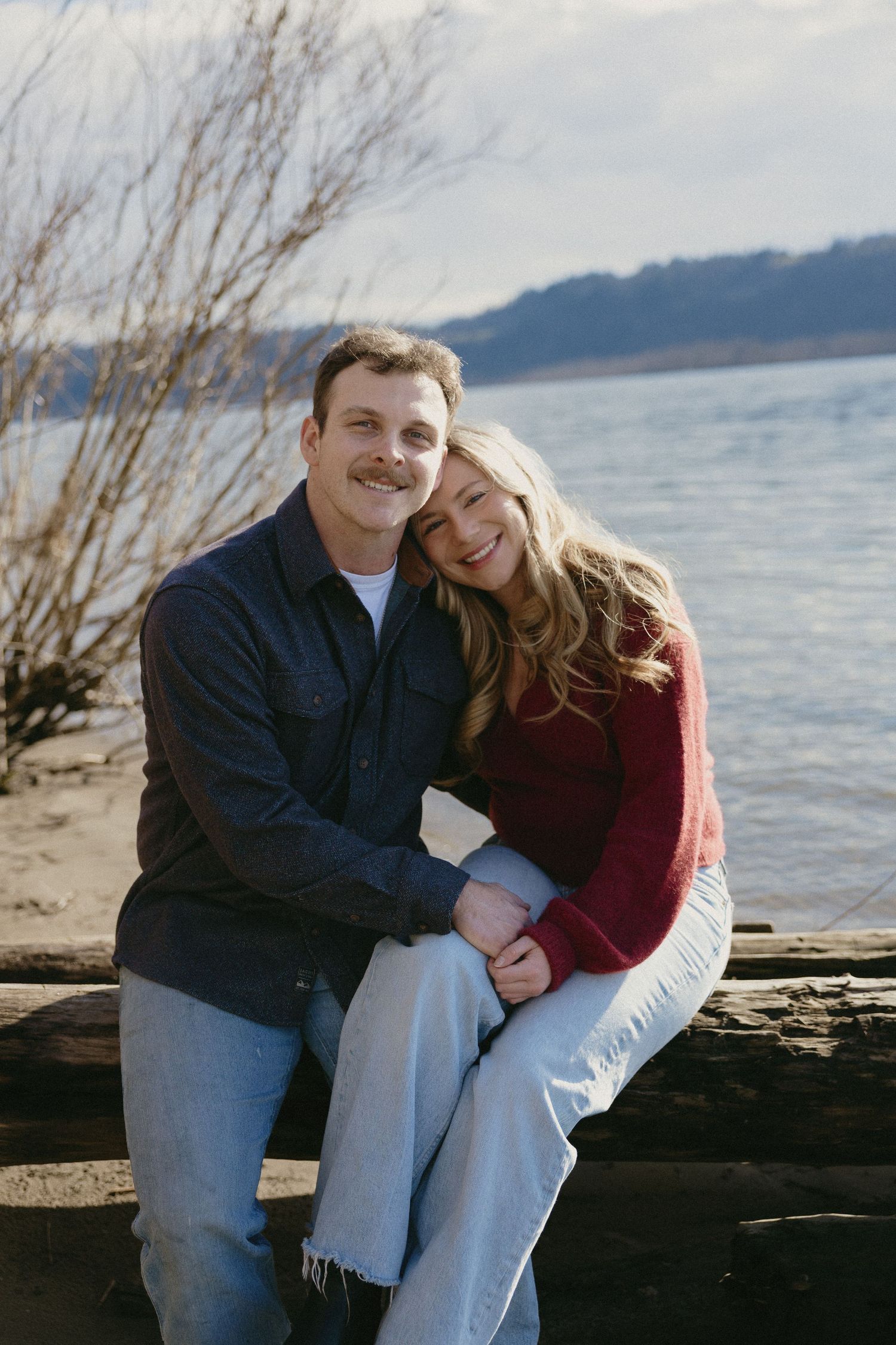 Smiling couple sitting together on a driftwood log near a lake shore on a sunny winter day.