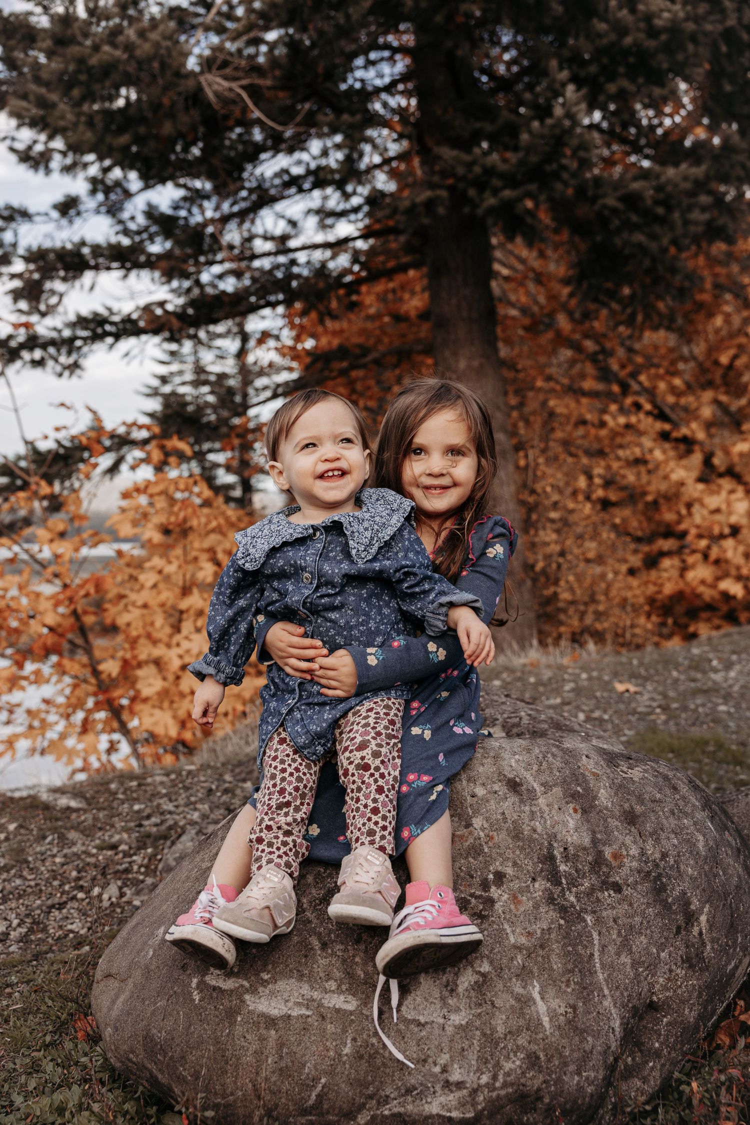 Two young sisters sitting on a rock surrounded by colorful autumn foliage smiling together.