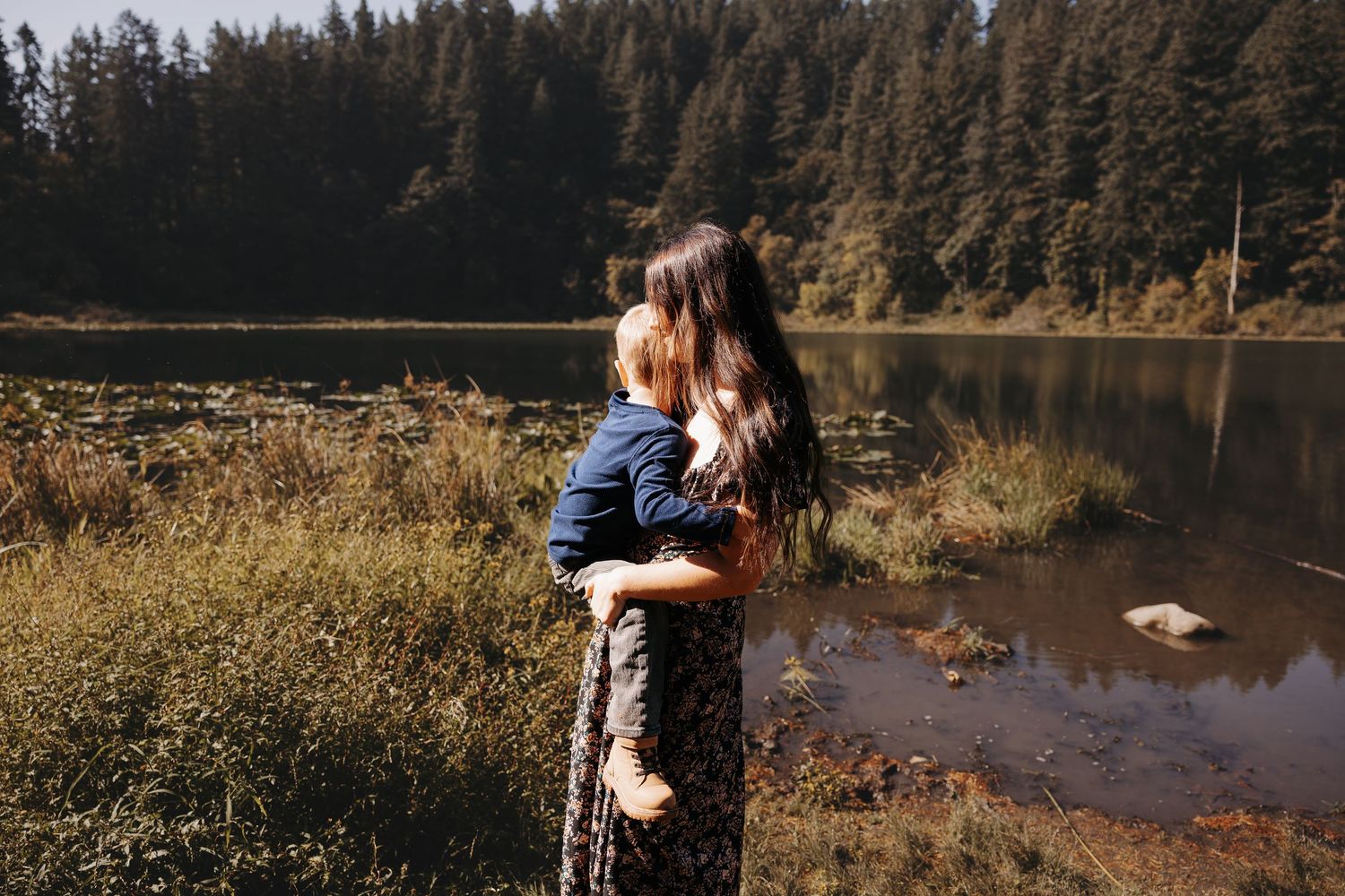 A mother holds her young child while standing by a serene forest lake surrounded by lush greenery.