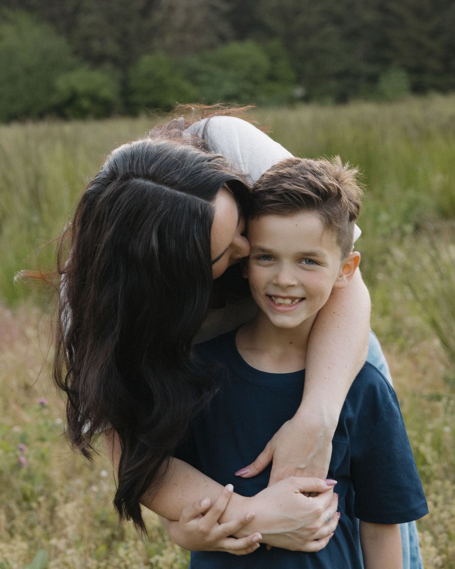 A woman with dark hair hugs and kisses a smiling young boy outdoors in a green meadow.