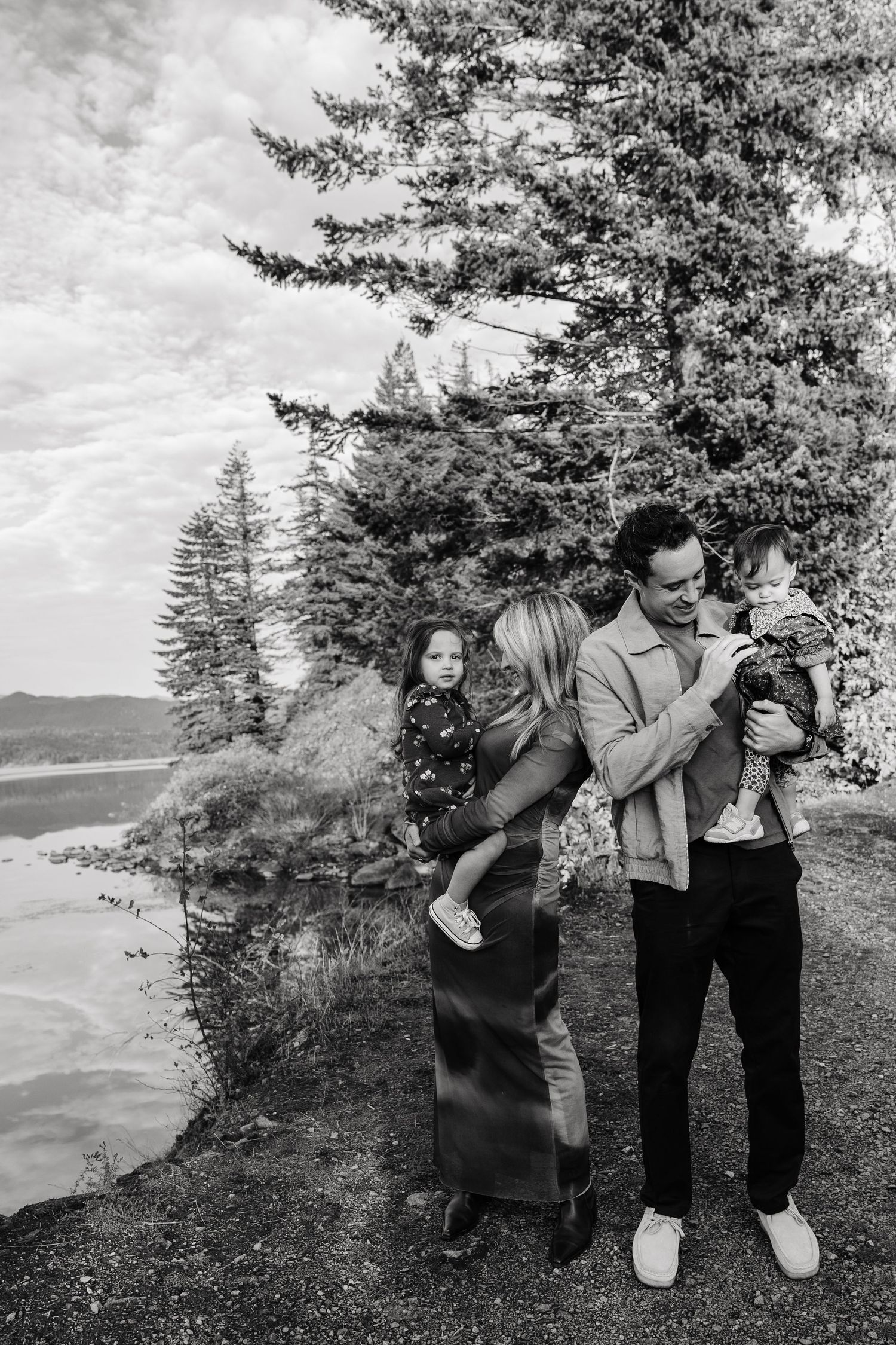 Black and white family photo with parents holding two young children near lakeside pine trees.