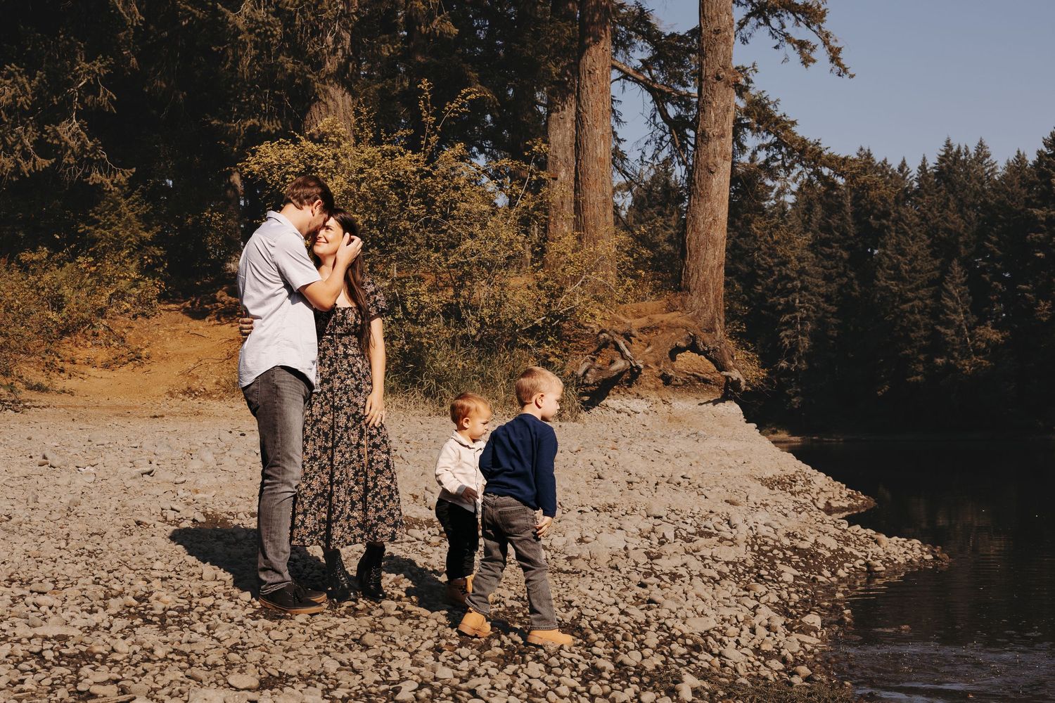 Family of four standing by rocky shoreline near tall trees, parents embracing with two boys.
