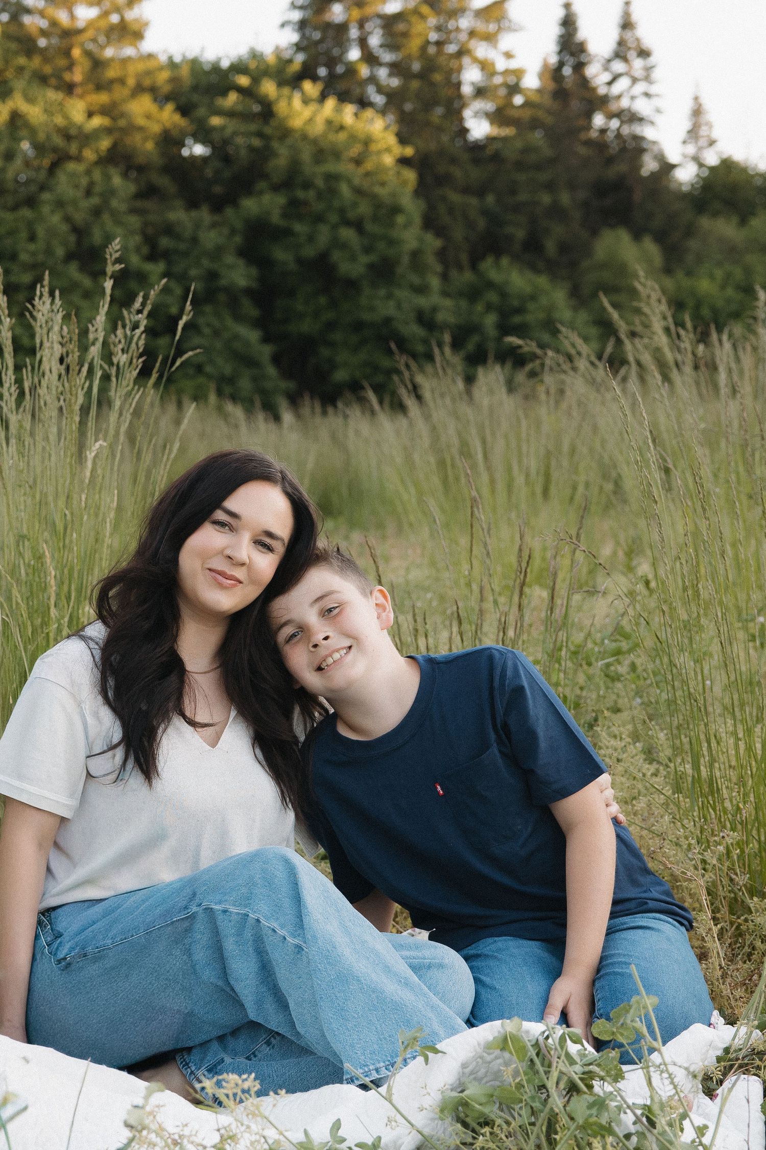 A dark-haired mother and her smiling son sit closely together on a blanket in a lush green field.