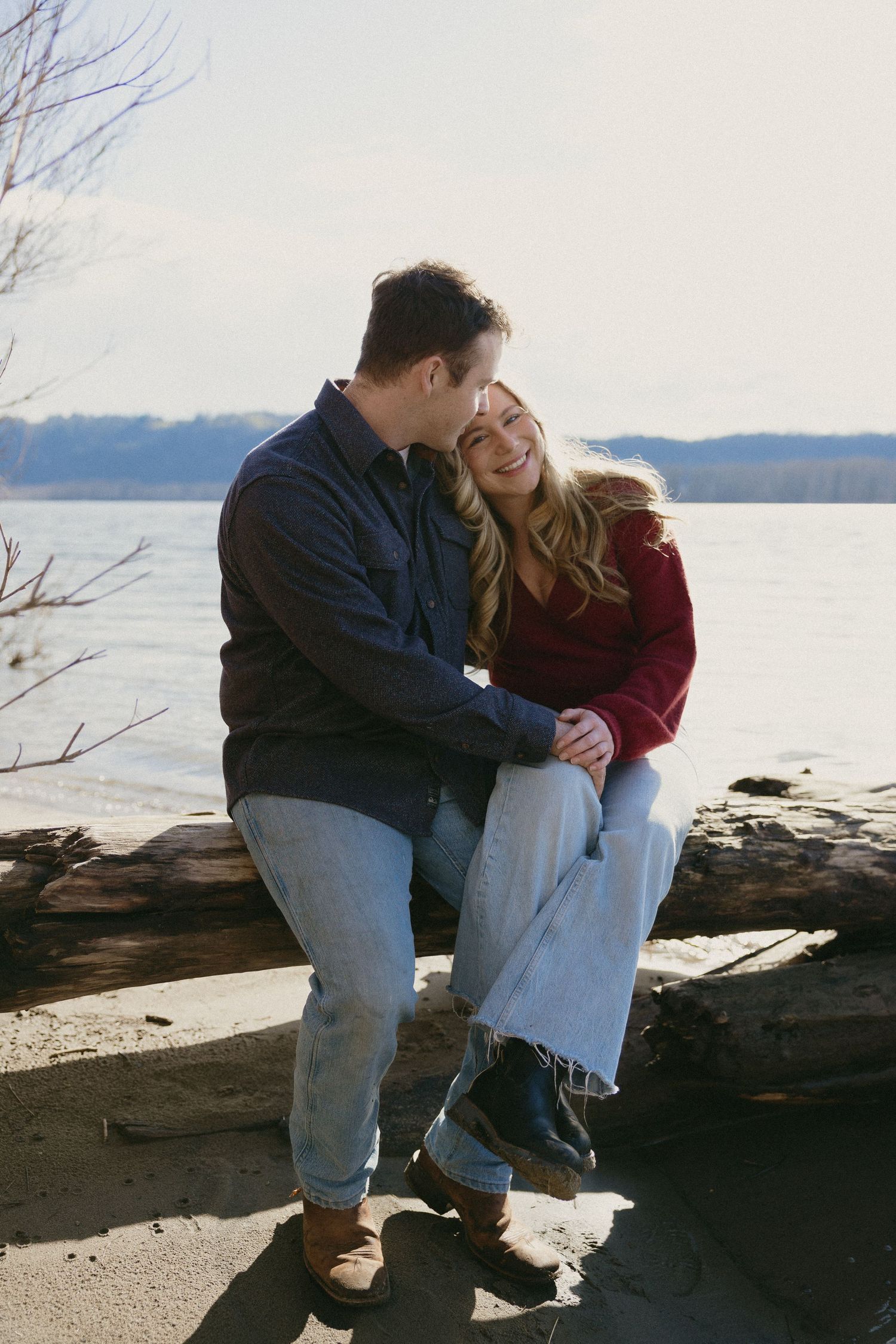 Happy couple sitting on a log by a lake, laughing and leaning close together in casual fall outfits.