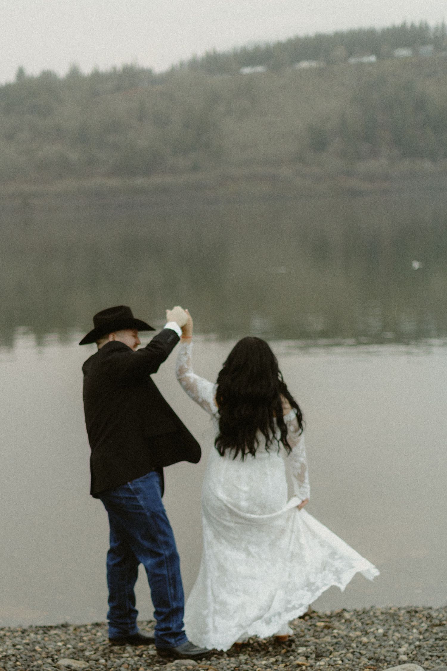 Bride and groom dancing by a misty lake, groom in black cowboy hat and suit, bride in white lace gown.