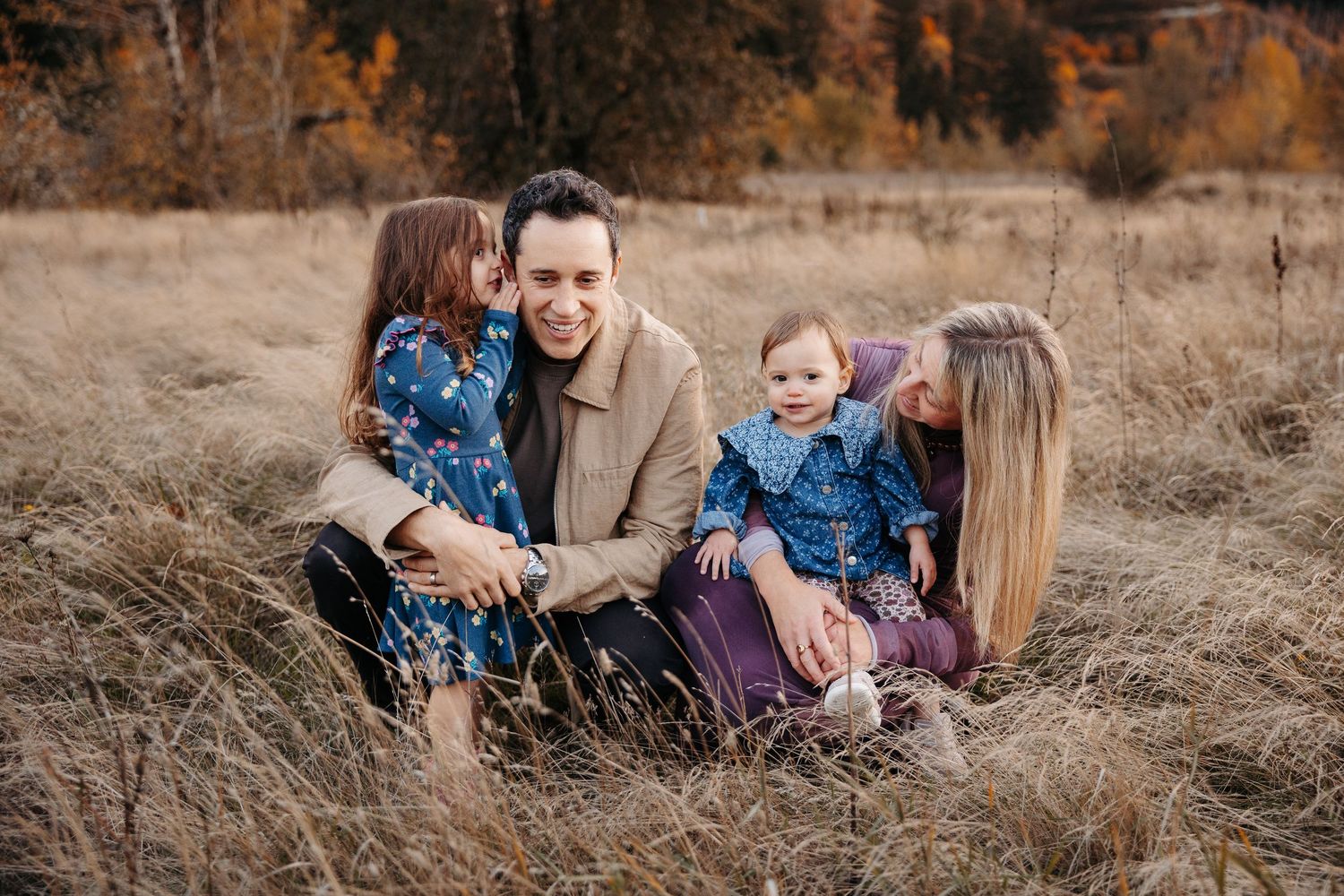 A joyful family of four sits together laughing in a golden autumn meadow.