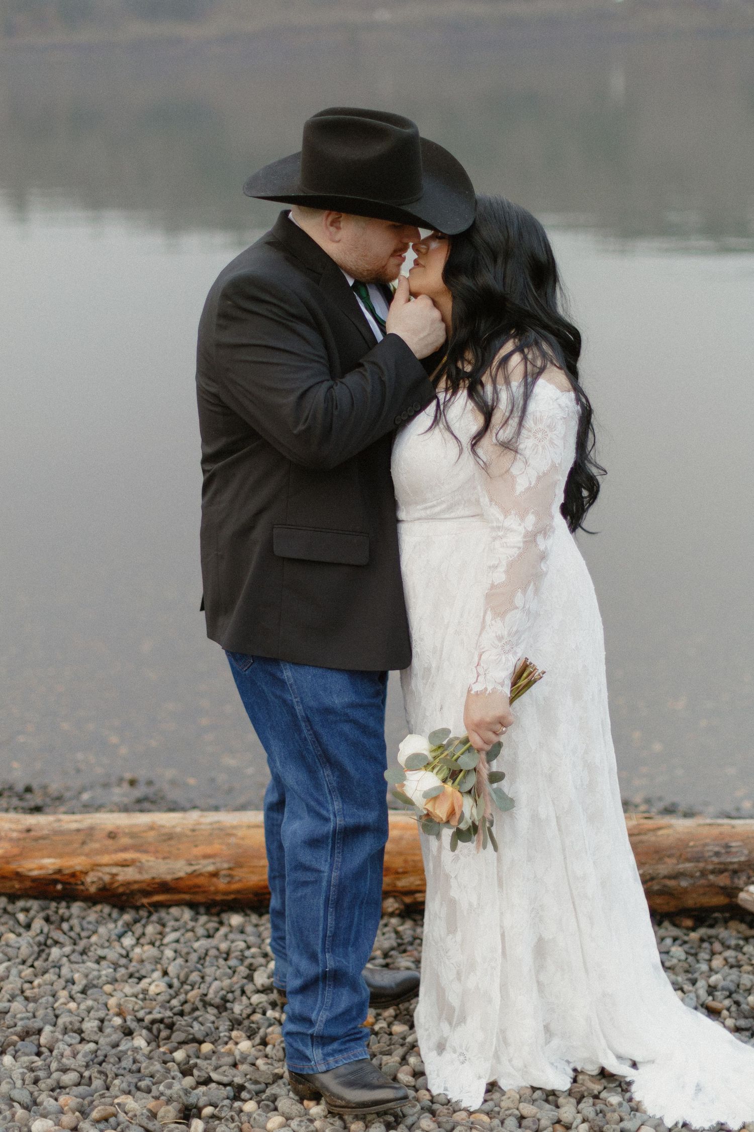 Couple shares a romantic kiss near a calm lake, bride in white lace dress holding a bouquet, groom in black cowboy hat.