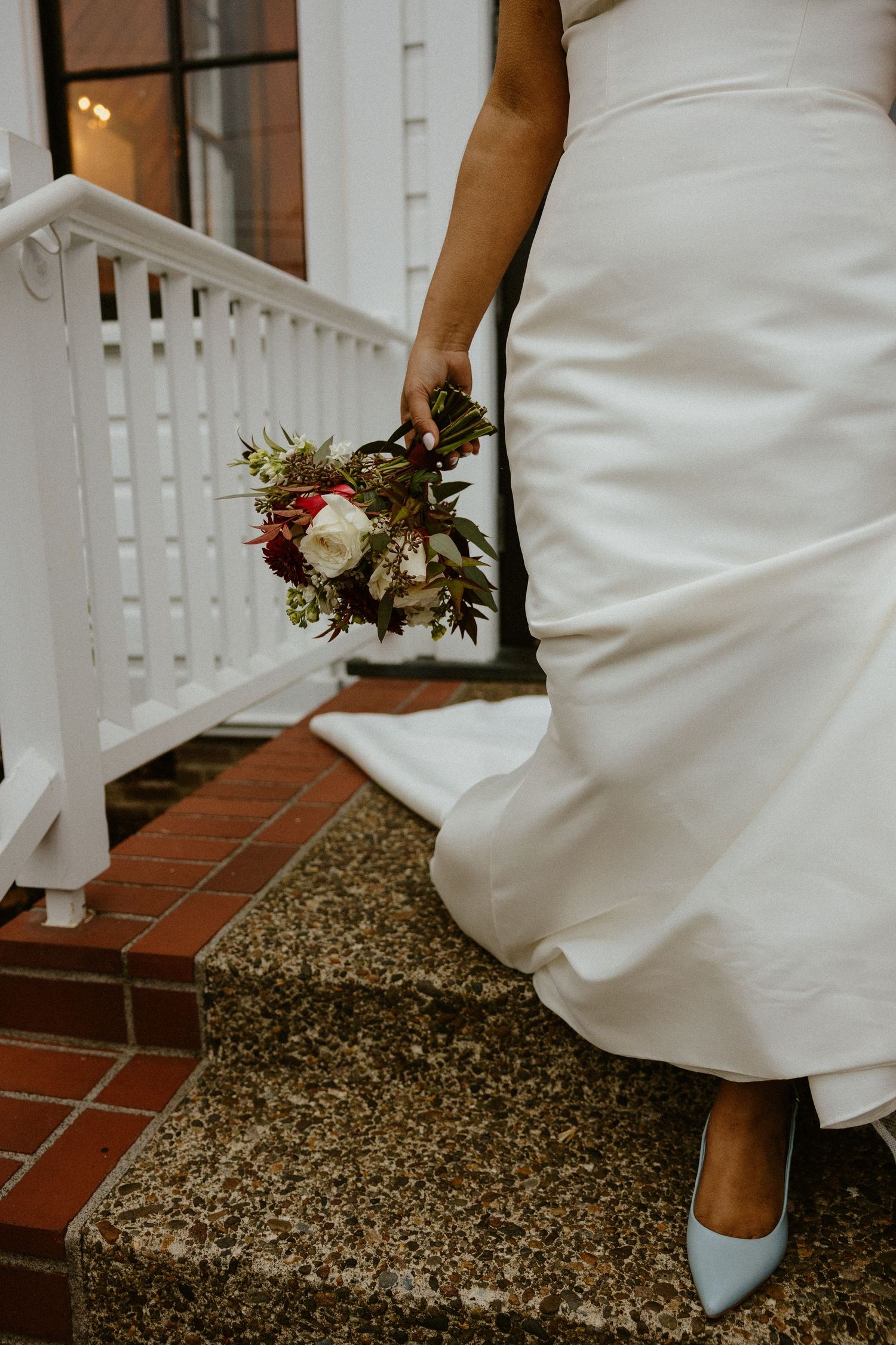 Bride in white gown holding red and white bouquet, wearing blue heels on stone steps.