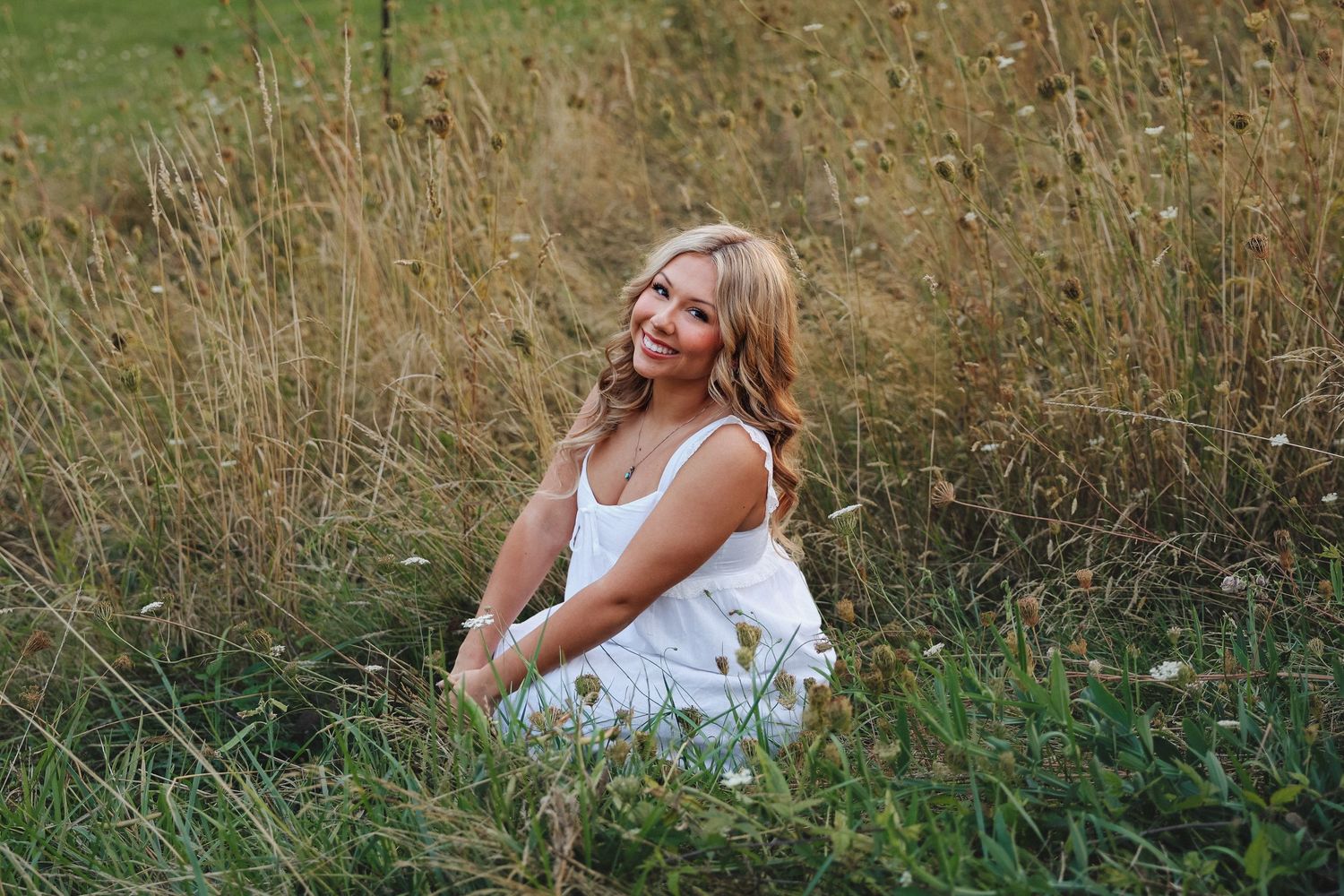 Smiling blonde woman in white dress sitting in tall grass field on sunny day.