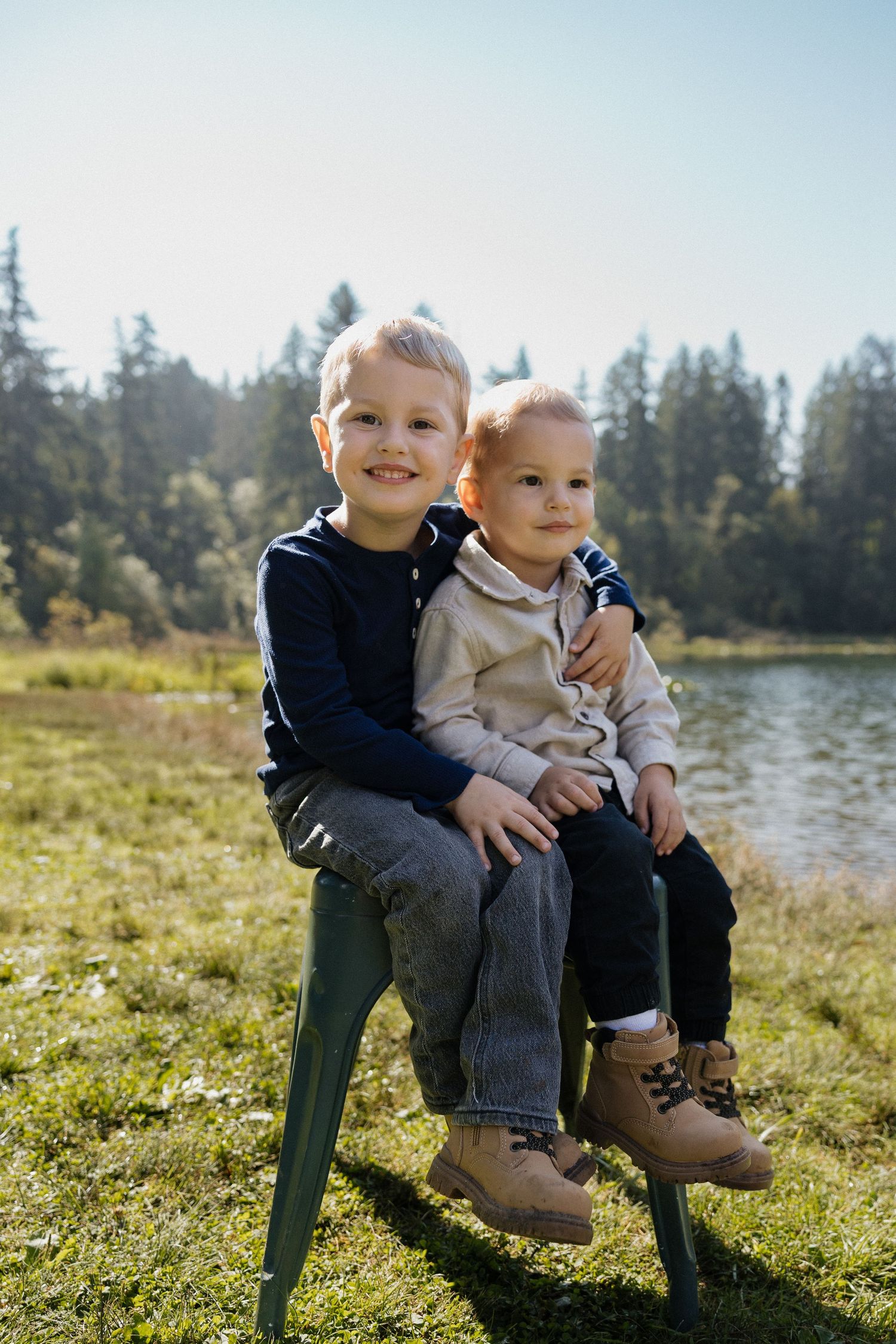 Two young boys sitting on a chair by a lake in a forest.